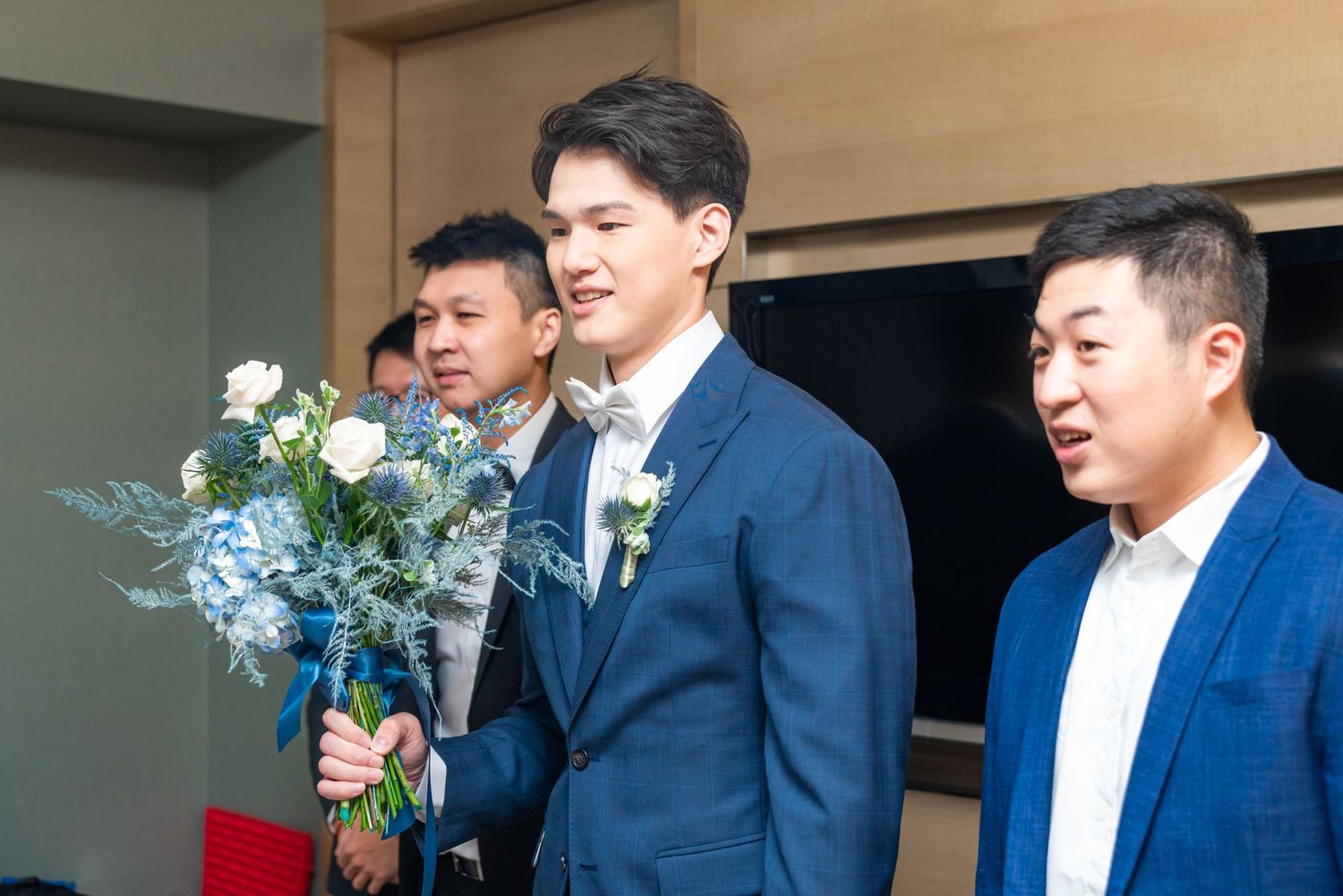 Three men in blue suits stand together at a formal event, with one holding a beautiful blue and white floral bouquet.