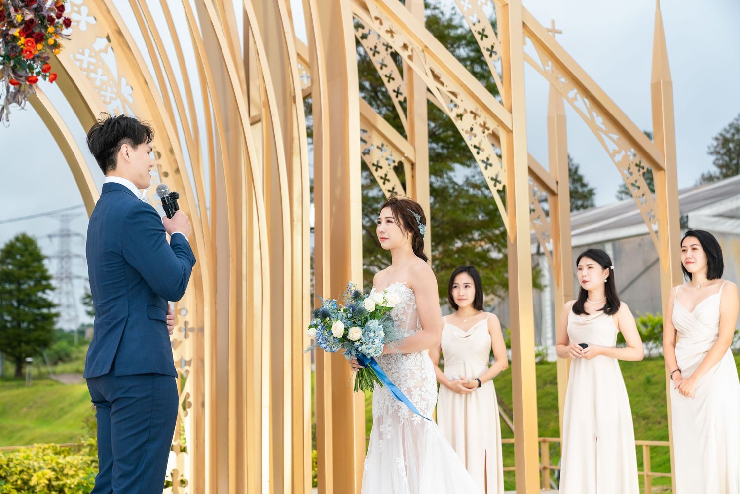 Outdoor wedding ceremony under modern wooden arbor with decorative beams against a green countryside landscape.