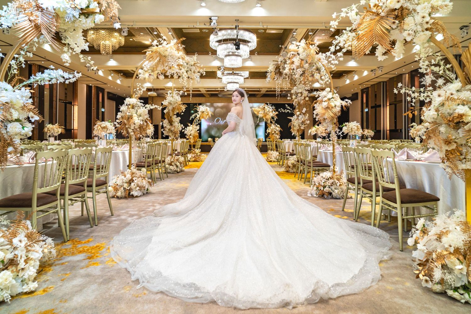 Luxurious wedding reception hall decorated with elaborate floral arrangements and a long flowing white wedding gown on display.