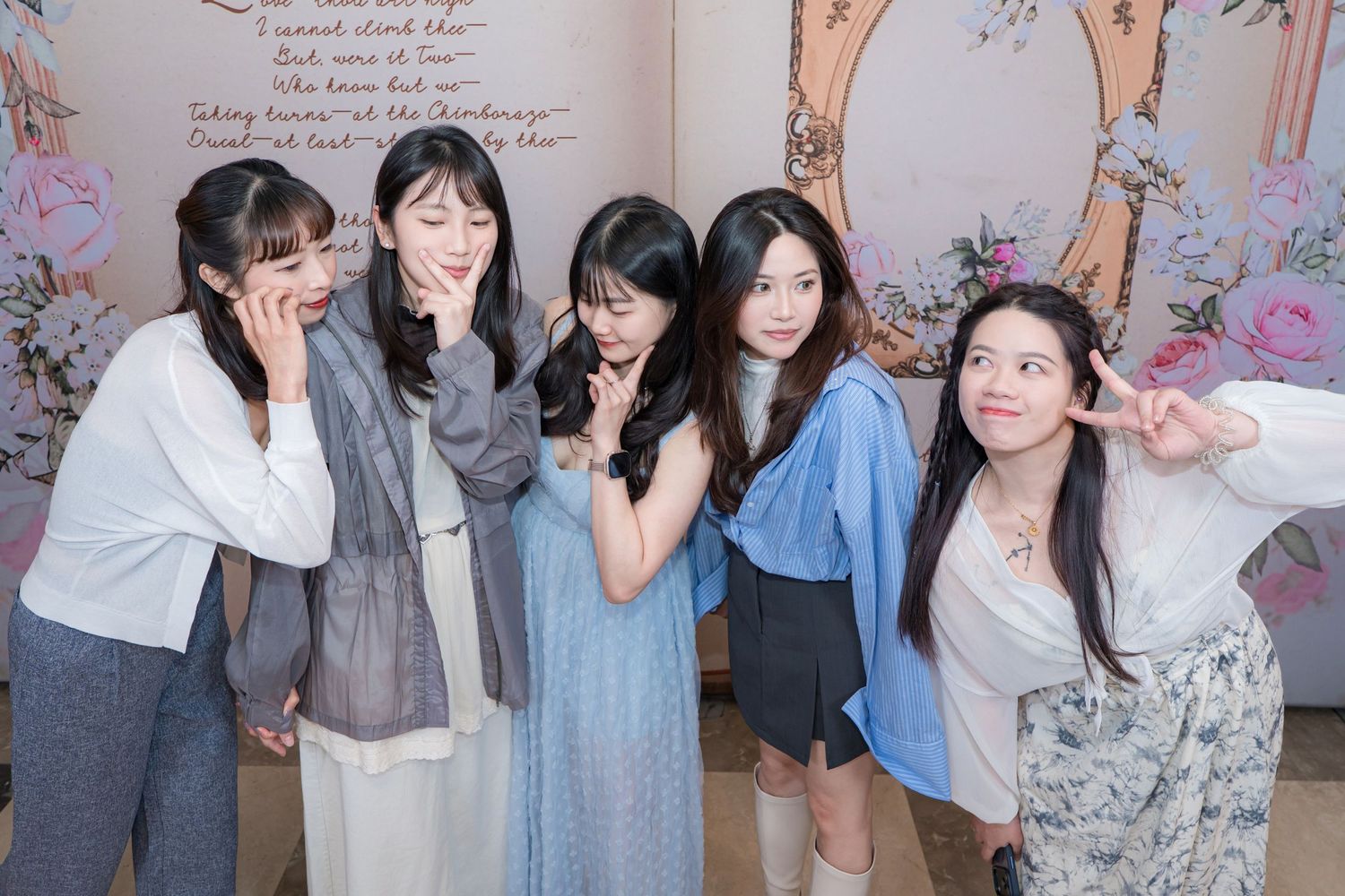 Five young Asian women posing together indoors in front of a floral decorated backdrop, smiling and making peace signs.