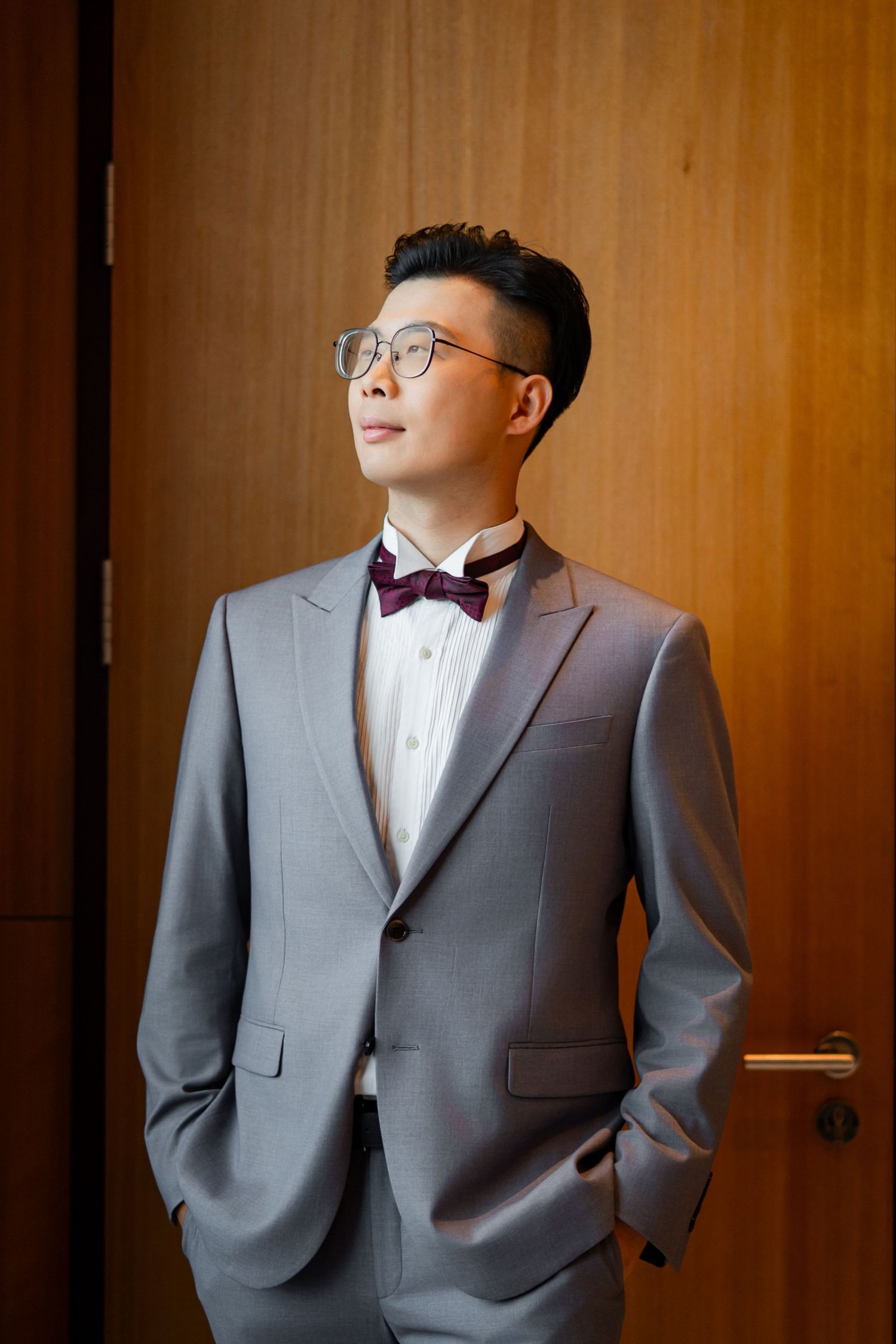 A groom in a grey suit with bow tie poses thoughtfully against a warm wooden wall.