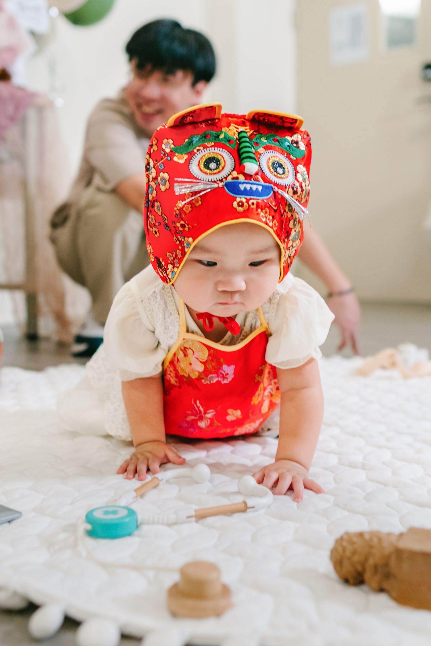 Baby wearing traditional red Chinese clothing crawls on white bedding while exploring toys and playthings.
