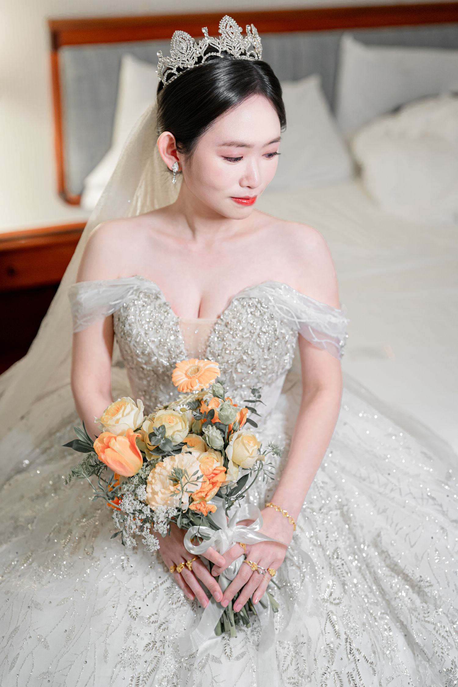 A bride in a sparkling beaded wedding gown holds a peach and cream rose bouquet in an elegant indoor setting.