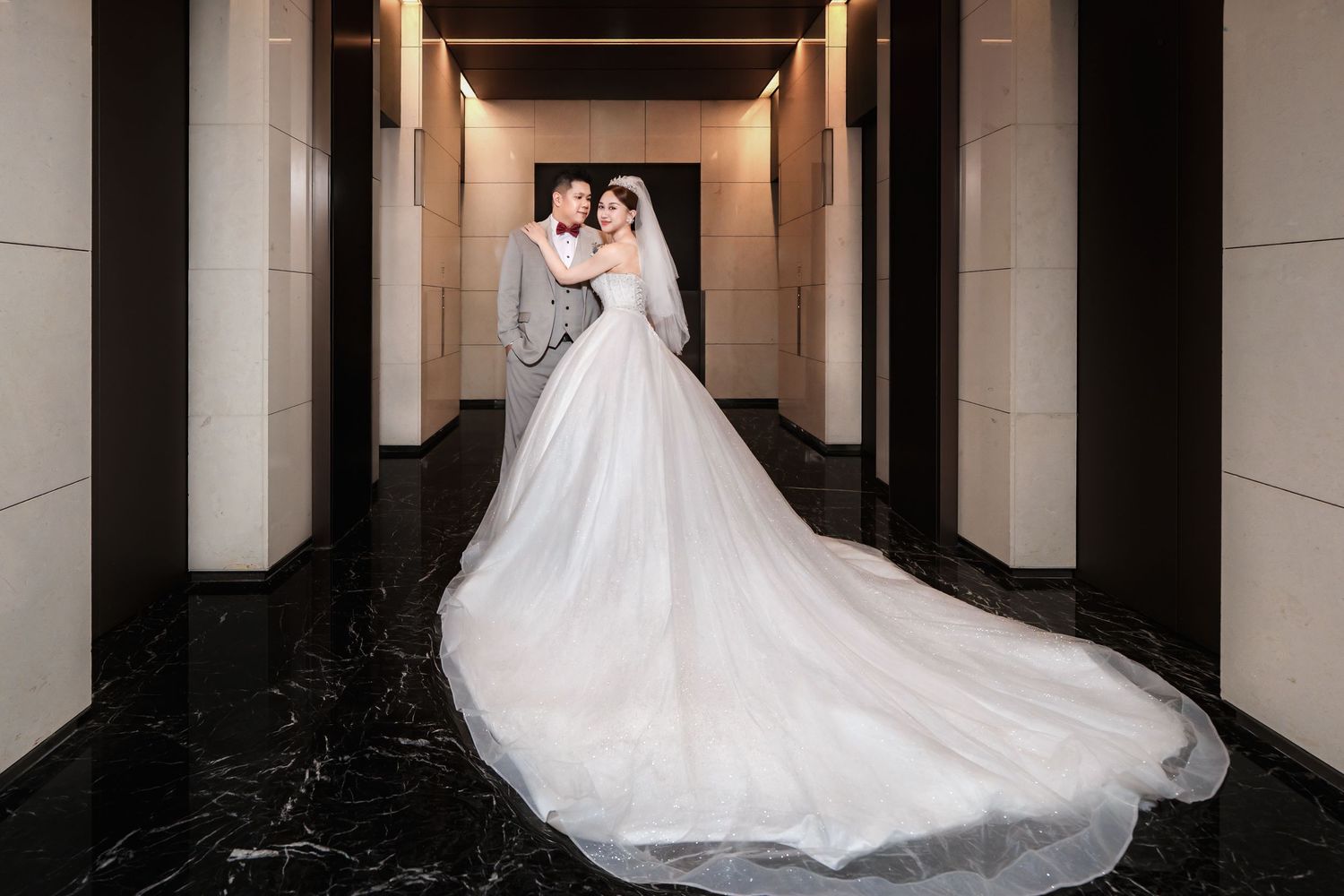 A bride in a flowing white wedding gown poses elegantly in a modern architectural hallway with dramatic lighting.