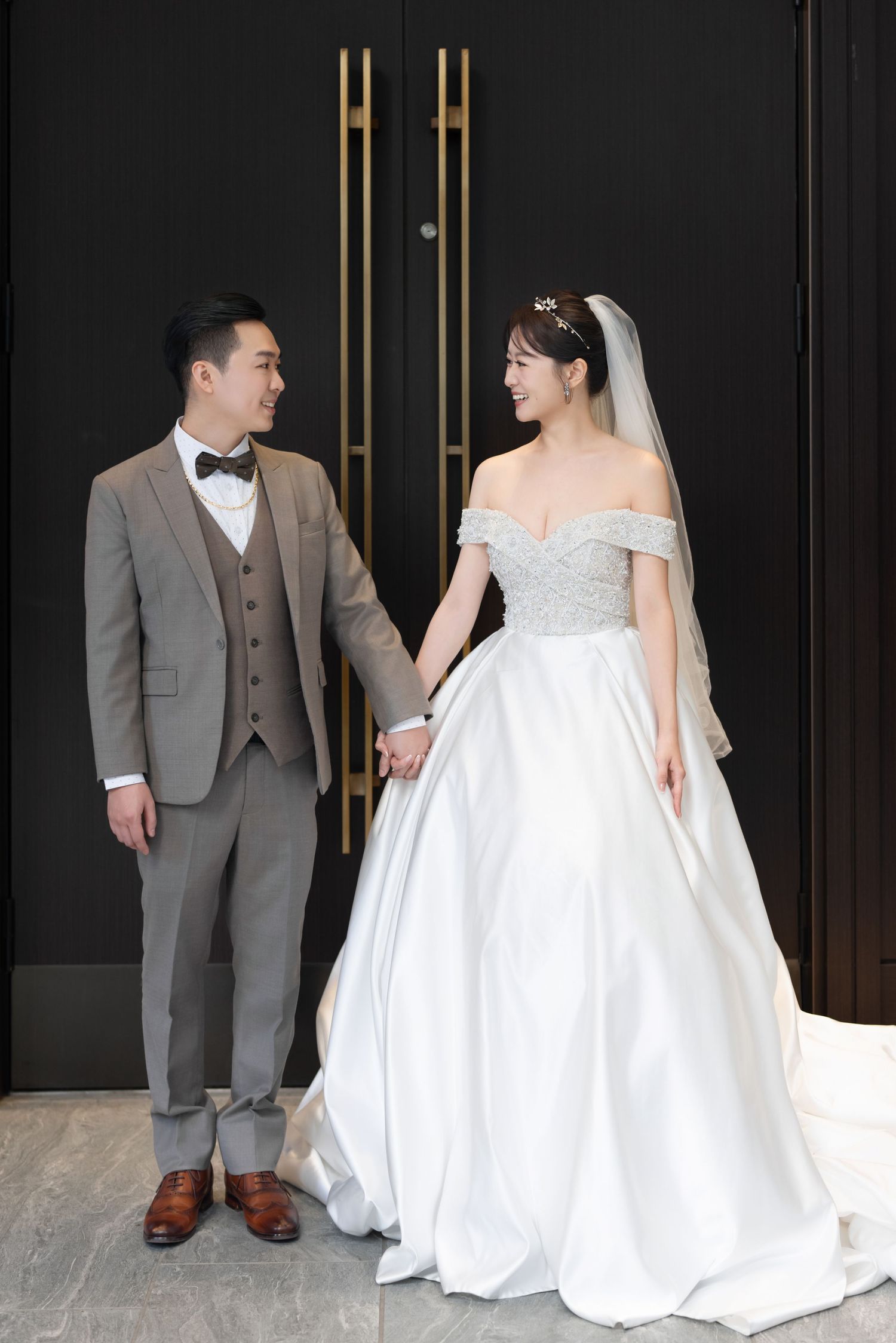 A couple in wedding attire stands together in front of a dark wall with gold accents.