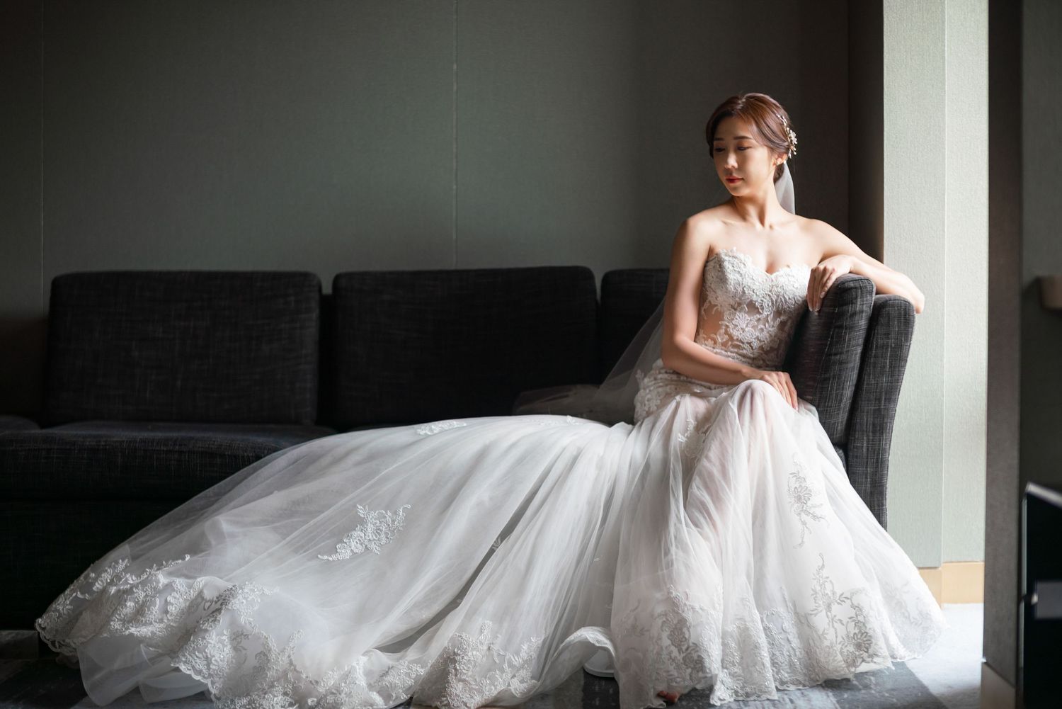 A bride in an elegant white lace wedding gown sits gracefully on a dark couch near a window in natural light.