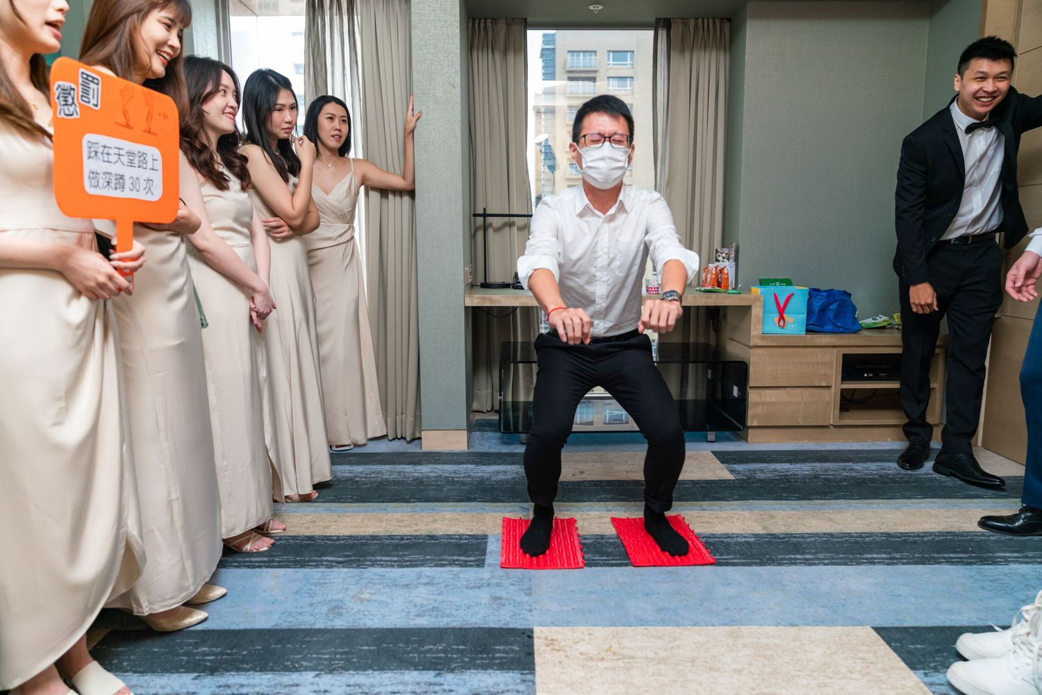 Wedding party members stand together in a hotel room while someone performs a traditional Chinese door game ceremony.