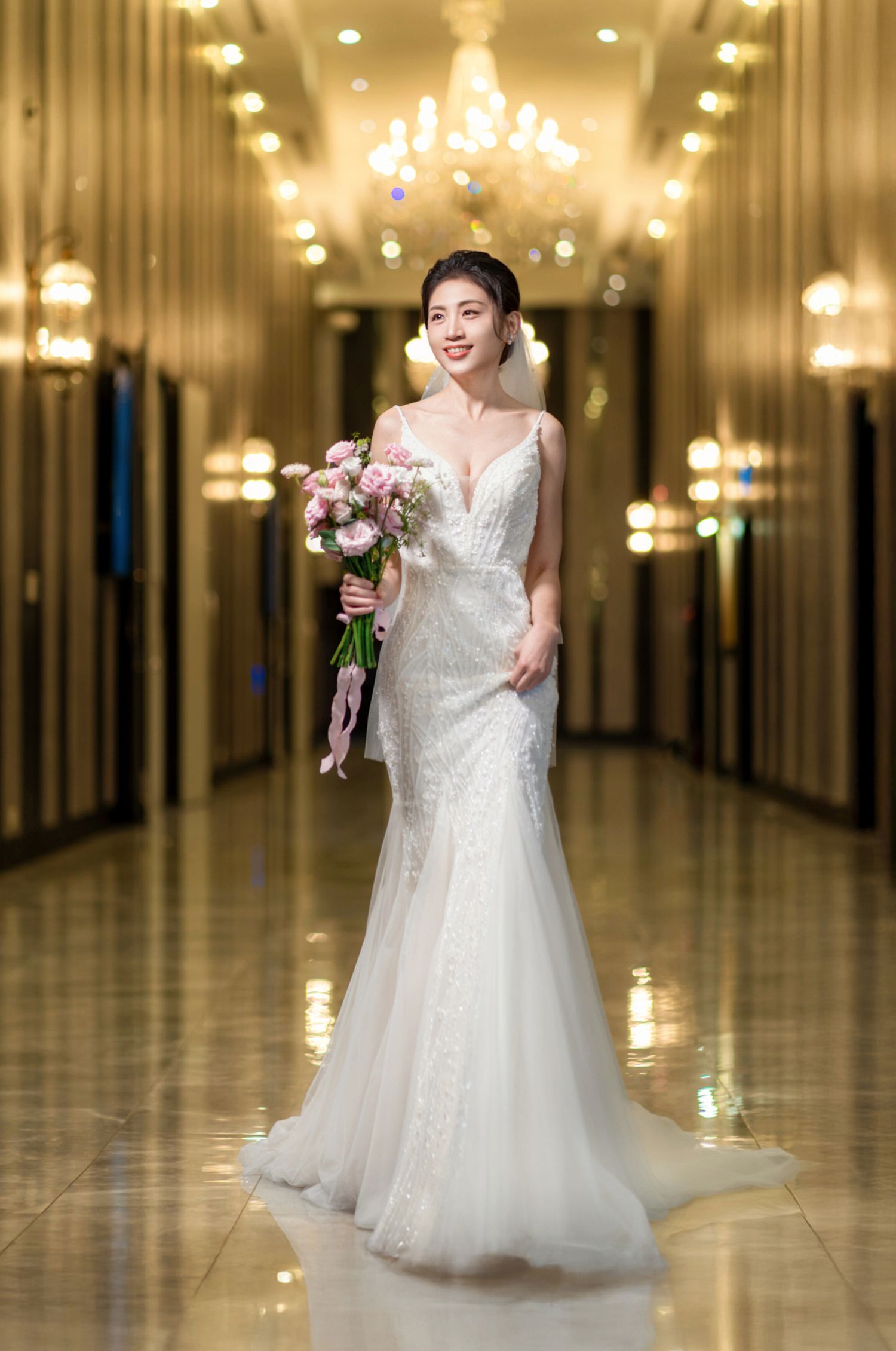 Elegant wedding gown with a fitted silhouette and floral bouquet captured in a grand hallway with crystal chandeliers.