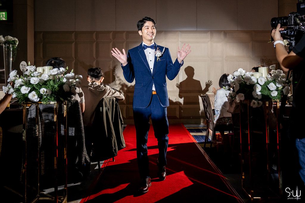 A speaker in a blue tuxedo gestures while standing on a red carpet with floral decorations.