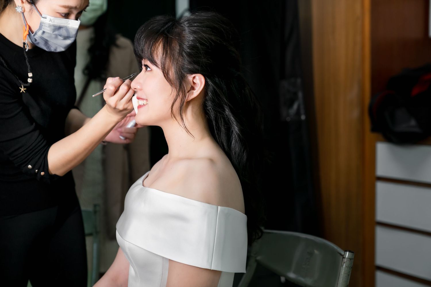 A bride in an off-shoulder wedding dress getting makeup applied by an artist wearing a mask.