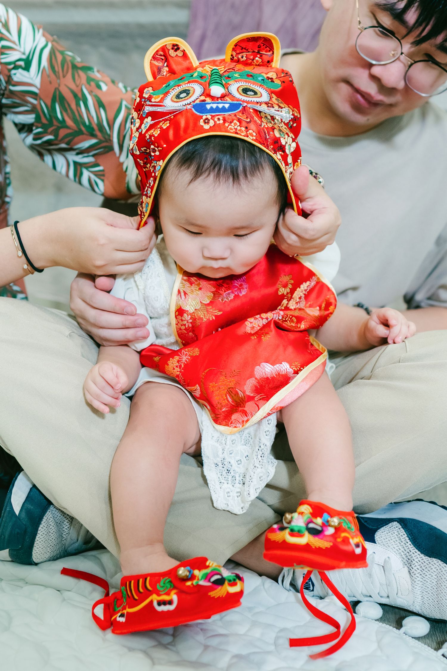 A baby wearing a traditional red Chinese dress and matching headband sits on a couch while being dressed.