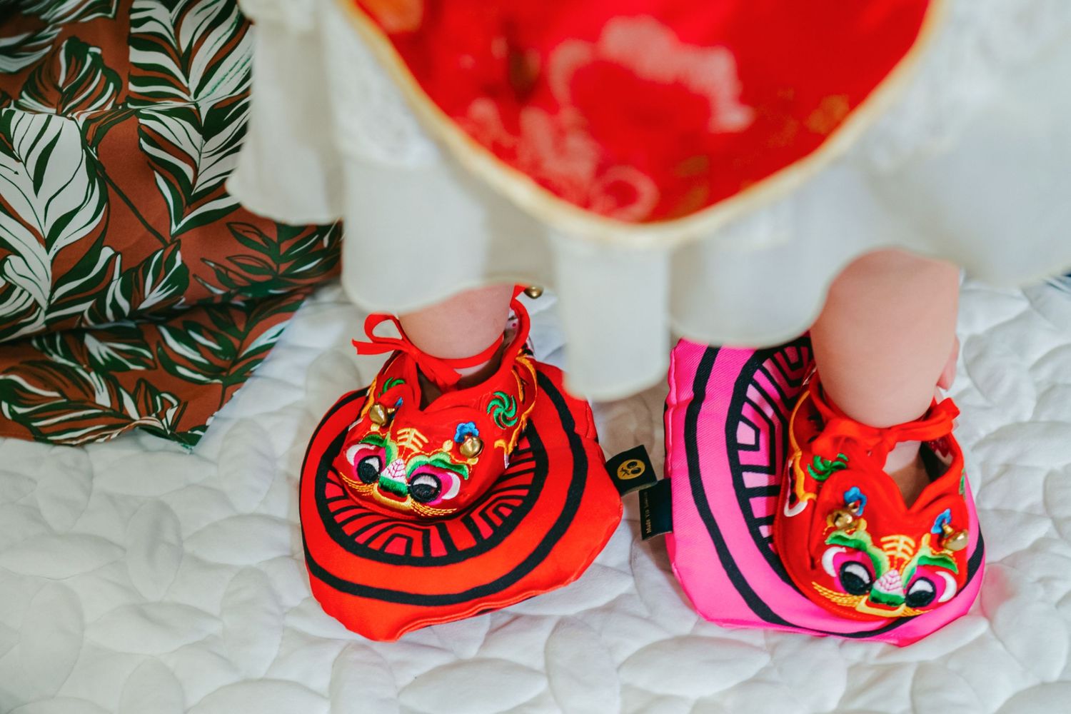 Traditional red and pink embroidered baby booties with fox designs displayed on white fabric with greenery.