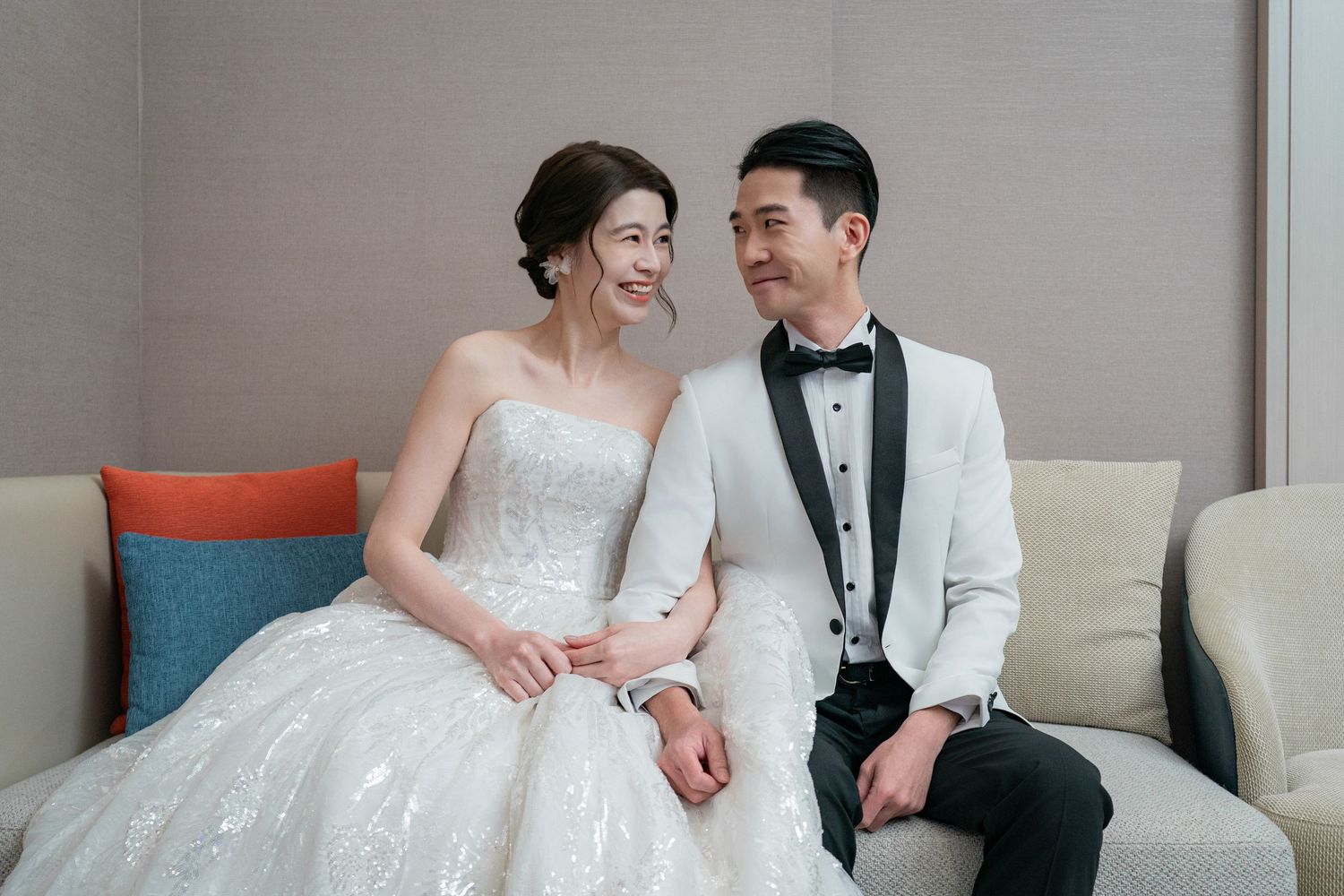 A happy bride and groom share a tender moment while sitting together on a couch in formal wedding attire.