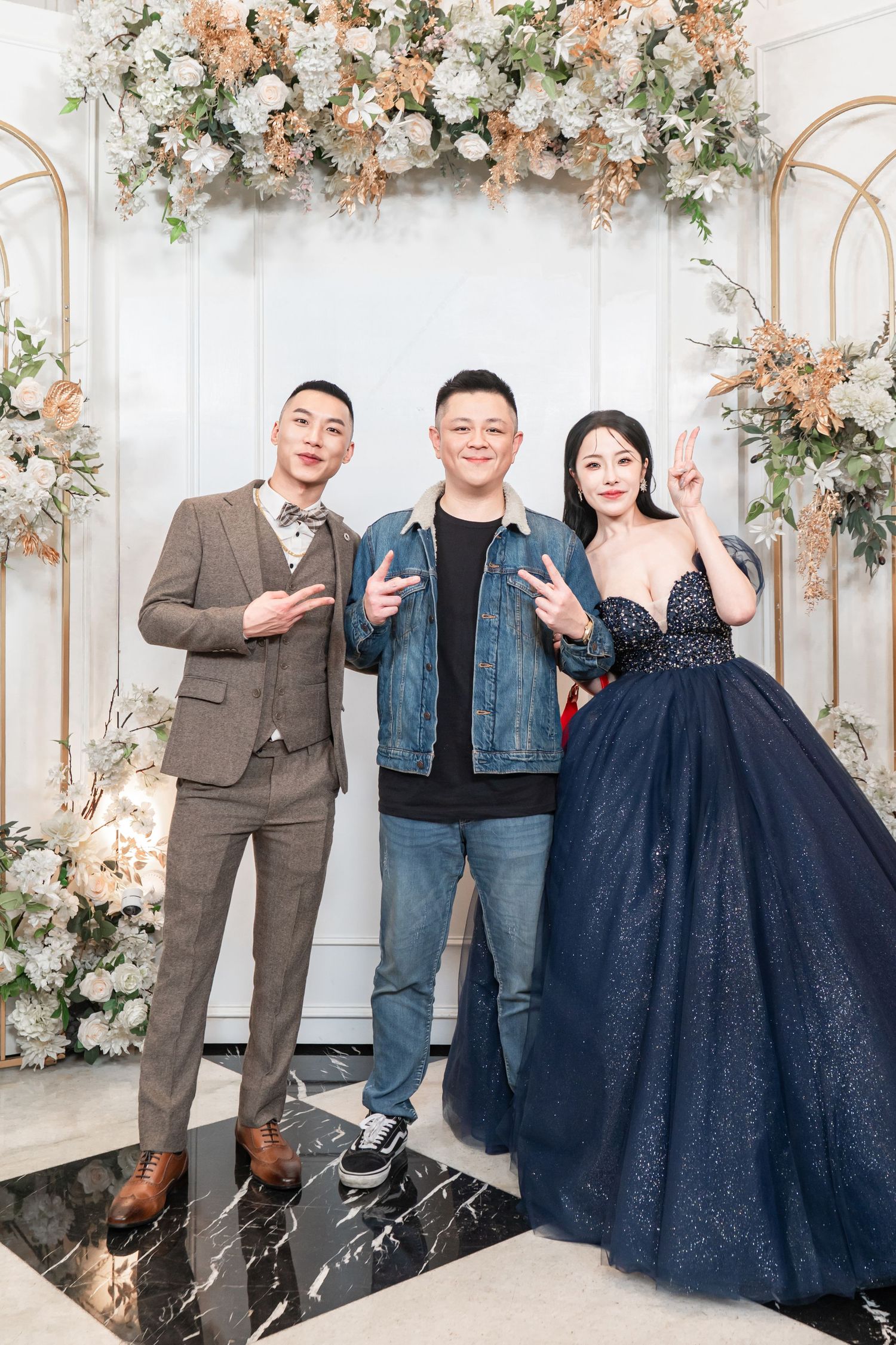 Three people pose in front of an elegant floral arch with white and peach blooms against a white backdrop.