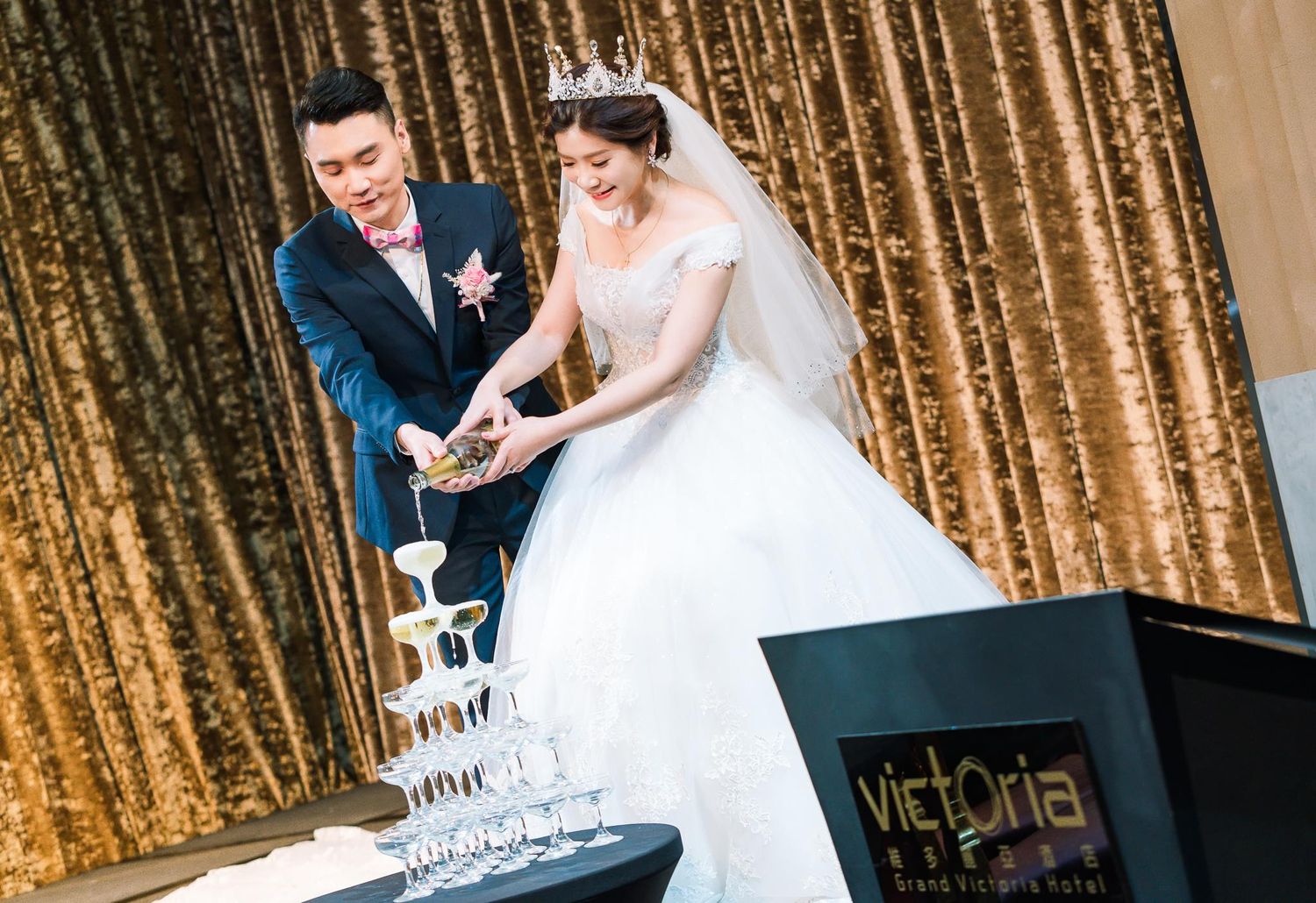 Newlyweds pour champagne into a tower of glasses against a shimmering gold backdrop at their wedding reception.