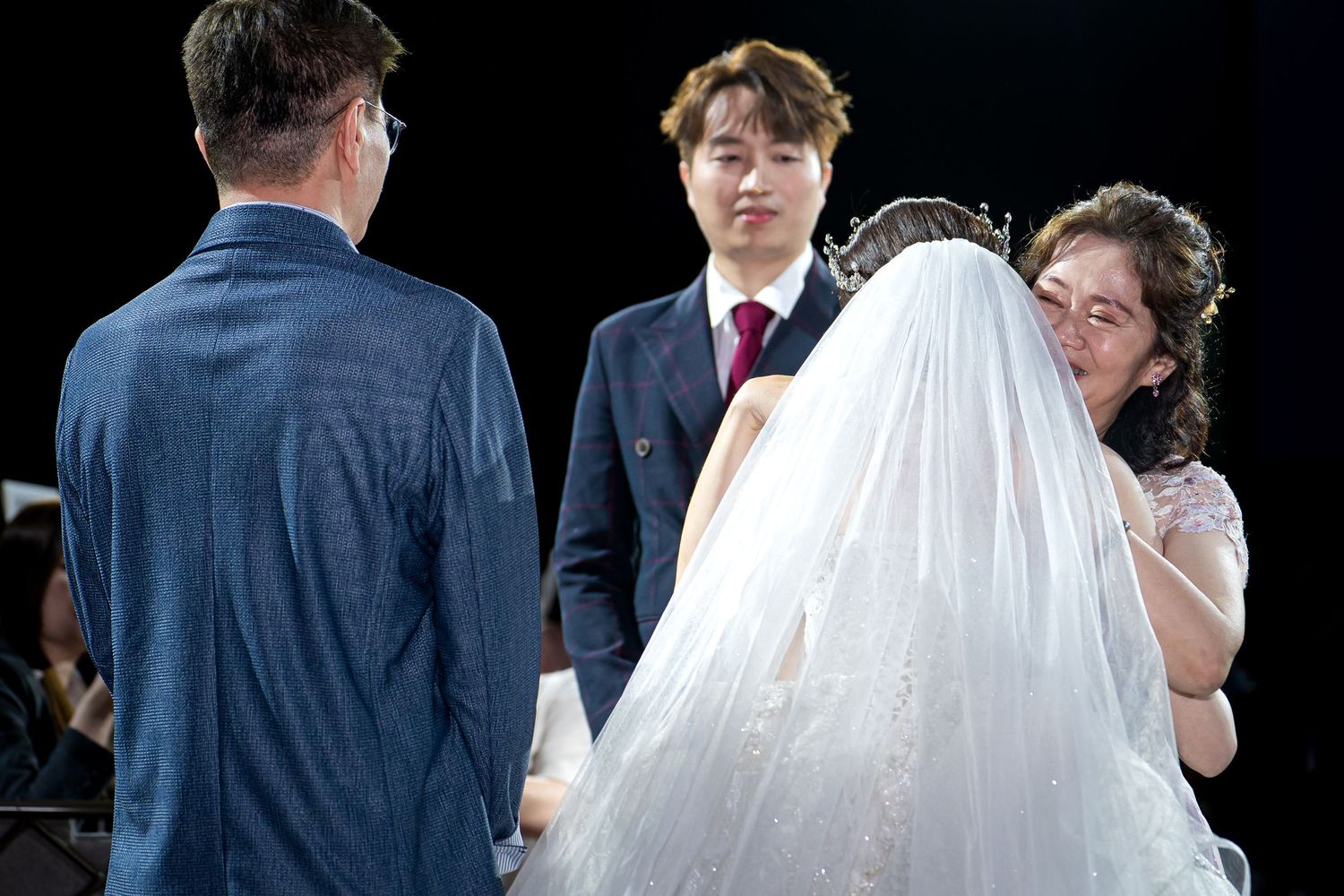 Wedding ceremony with bride in white veil, groom in suit, officiant, and guest smiling on stage.