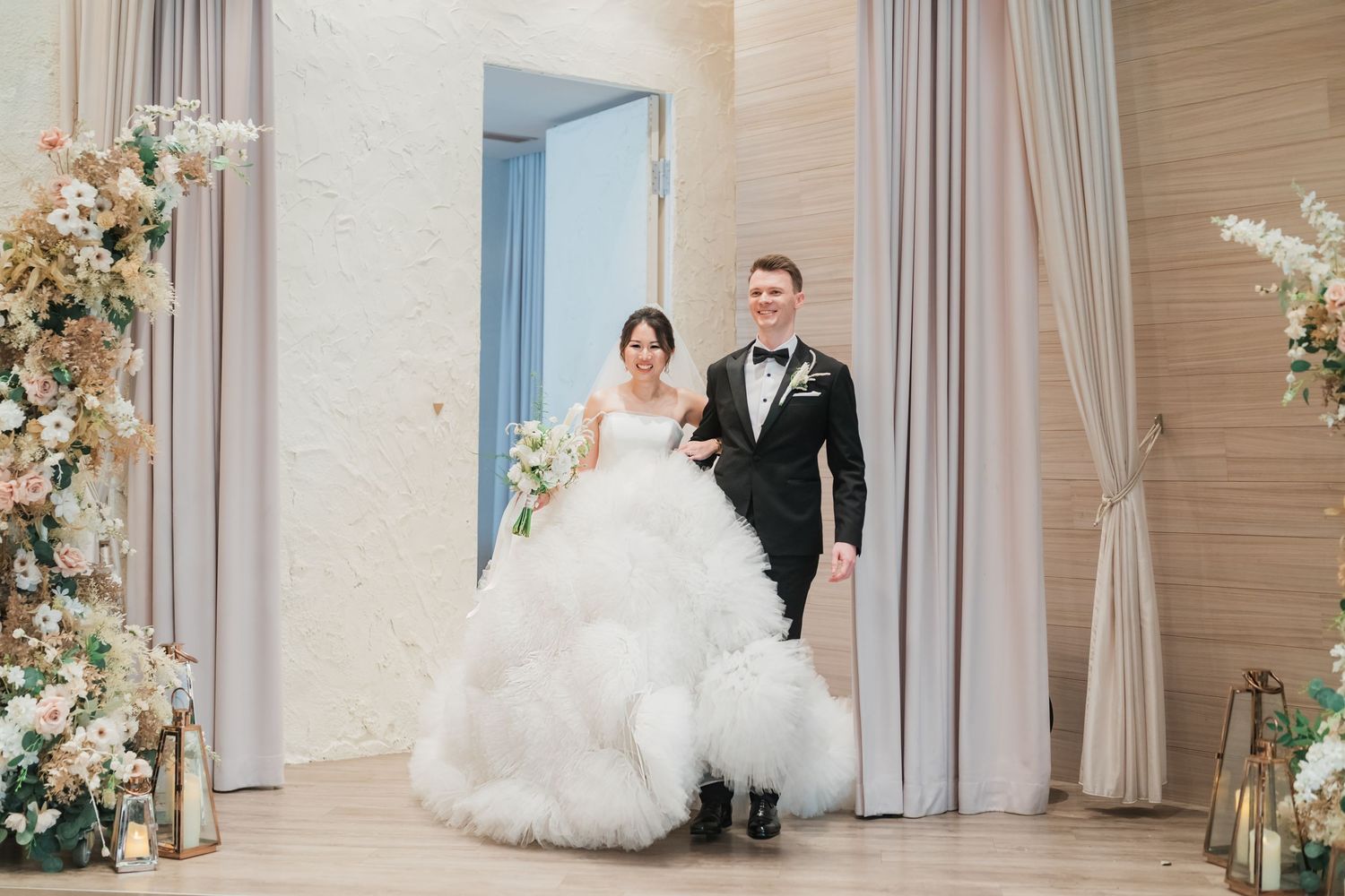 A bride and groom dance together in an elegant ballroom decorated with floral arrangements and soft curtains.