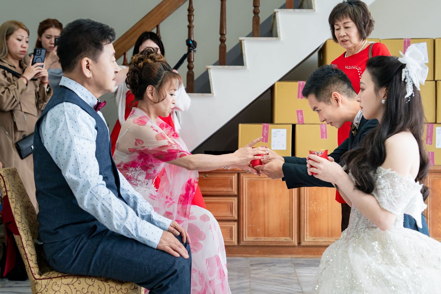 Traditional Chinese wedding tea ceremony with bride in white gown and guests exchanging red envelopes indoors.