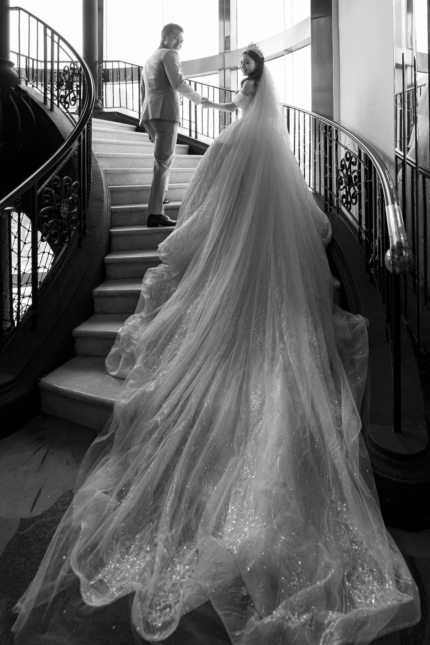 Black and white photo of bride in long flowing veil and gown ascending elegant spiral staircase with groom.
