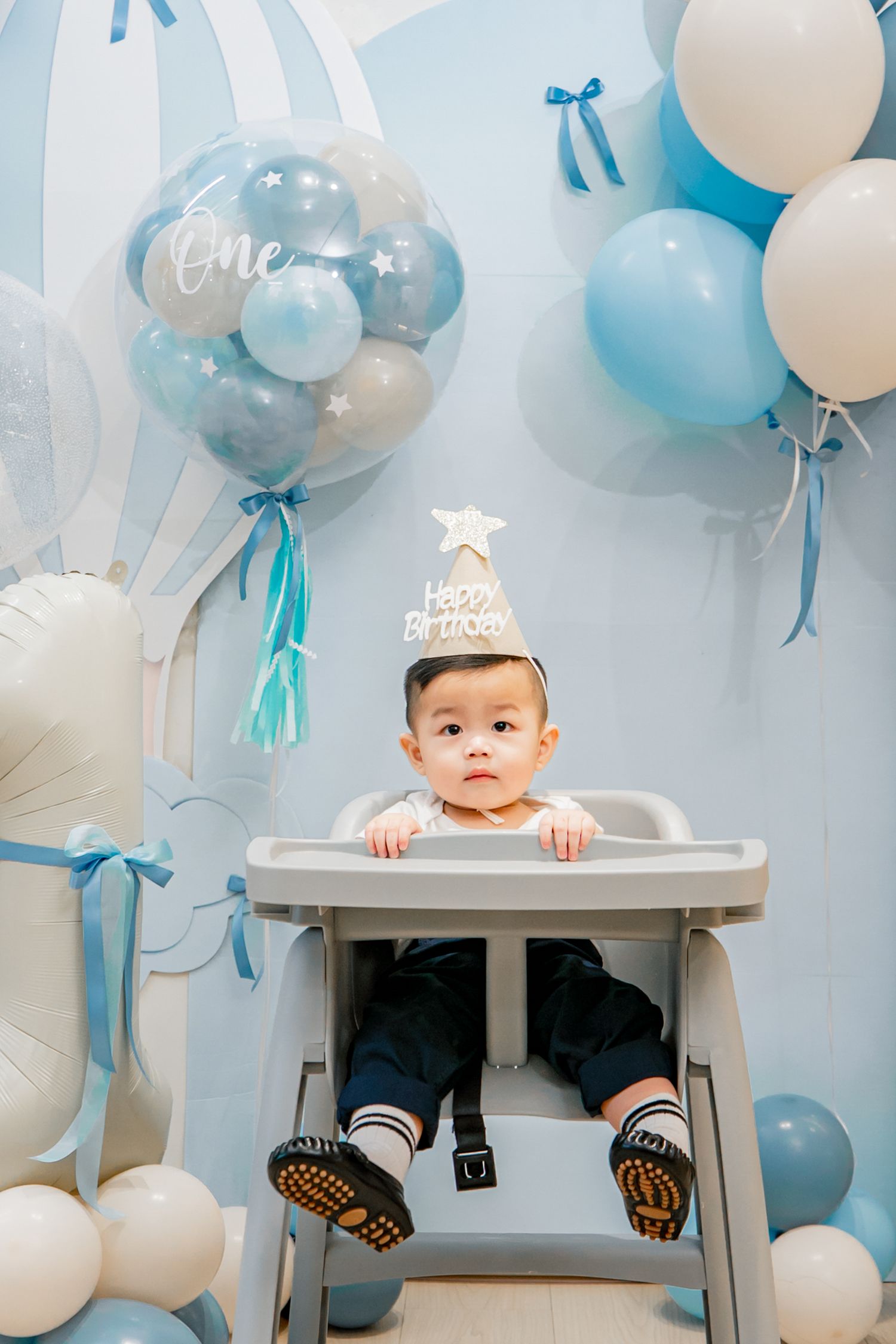 Baby in high chair celebrates first birthday with blue and white balloon decorations in background.