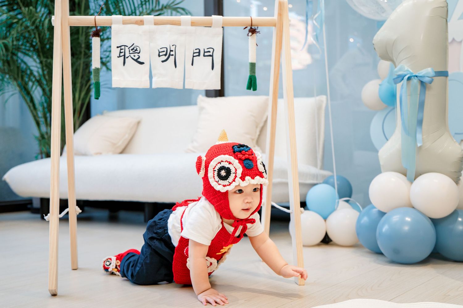 Baby in red onesie crawls across white floor with blue and white balloons in decorated room.