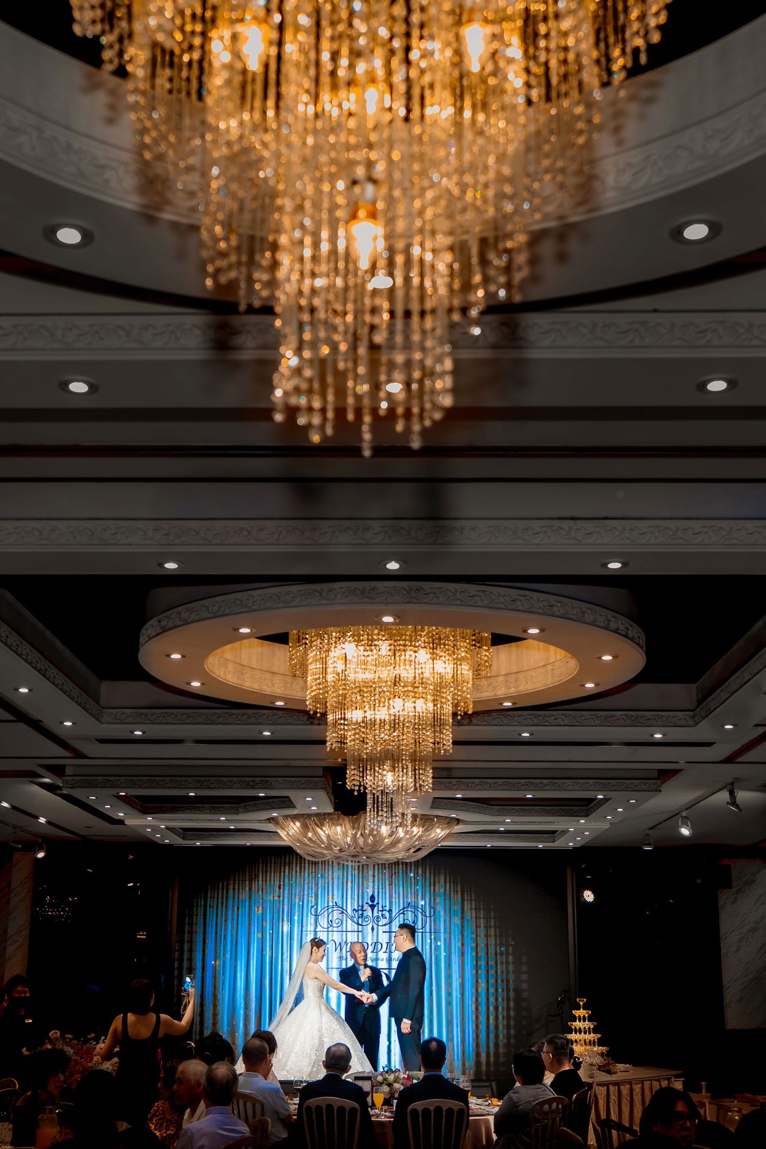 Elegant wedding ceremony setup in a ballroom with crystal chandeliers and blue accent lighting on stage.
