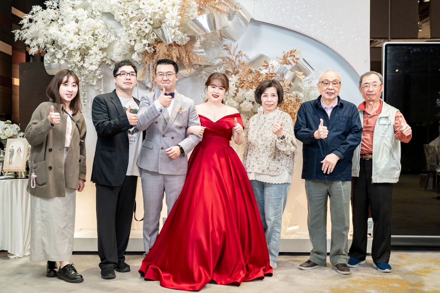 Wedding party in formal attire with bridesmaids in red dresses and groomsmen in gray suits pose together for photos.