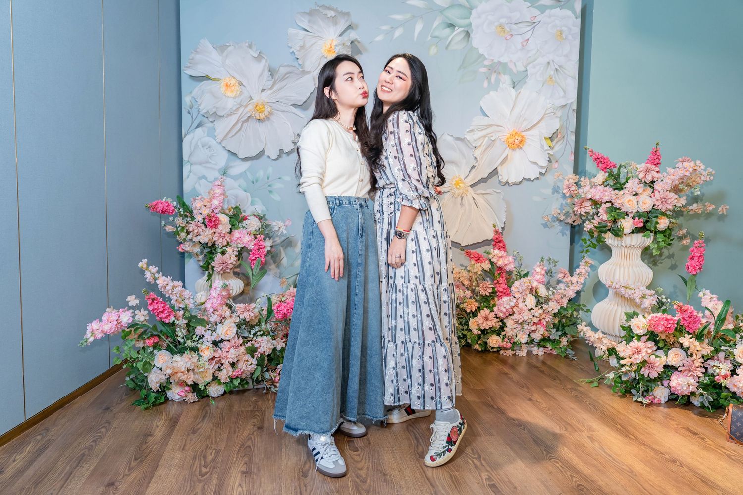 Two people pose together against a floral backdrop with pink flowers and a blue wall in a studio setting.