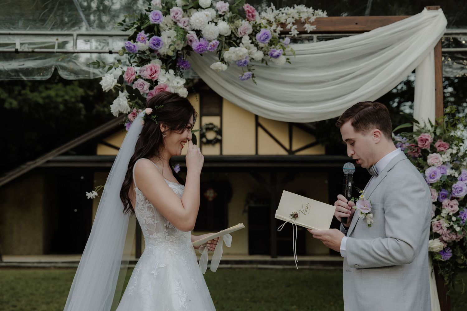 Bride and groom exchanging vows outdoors under a floral arch adorned with pink and white flowers at a wedding ceremony.