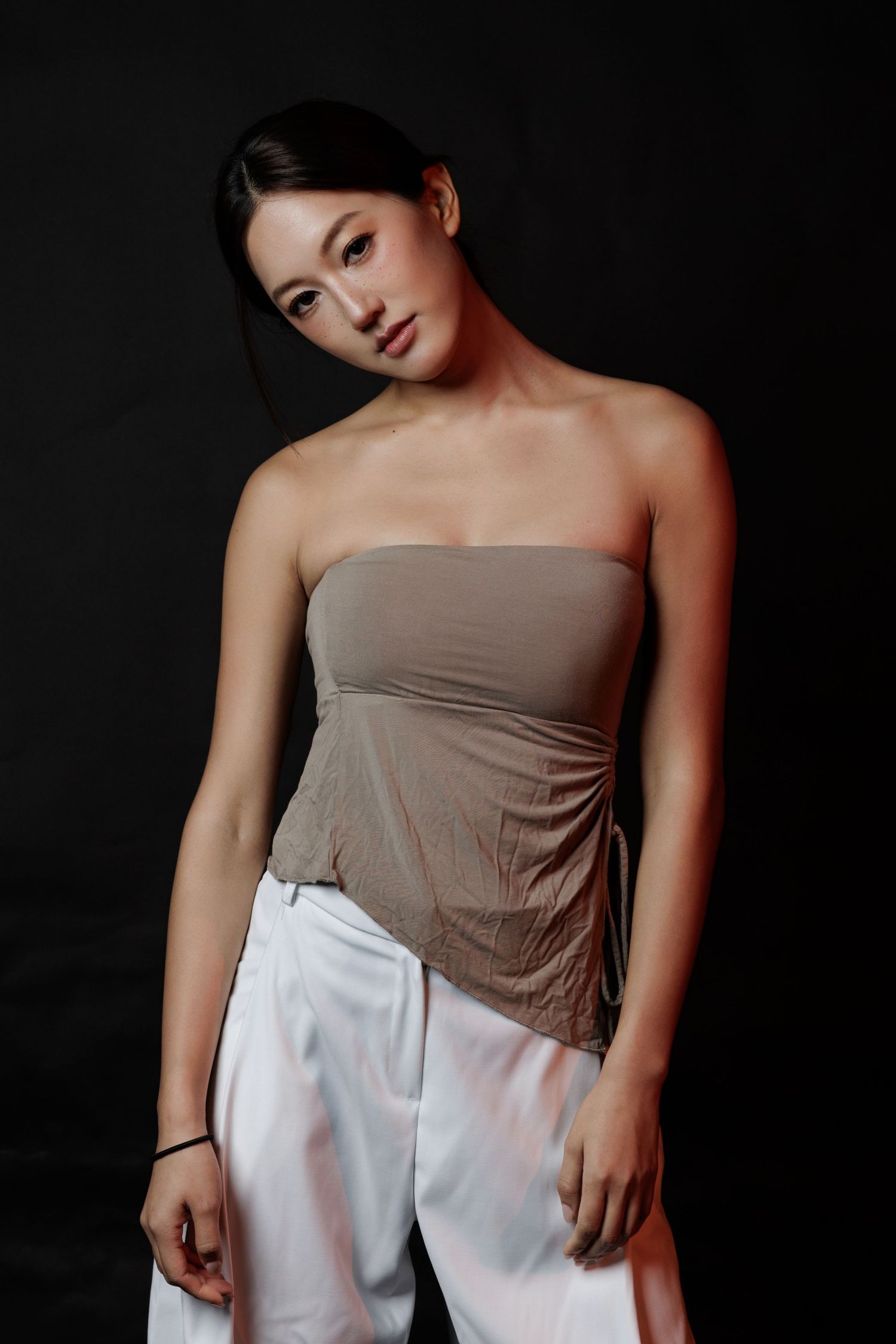 Young woman in strapless brown top and white pants posing against dark background.