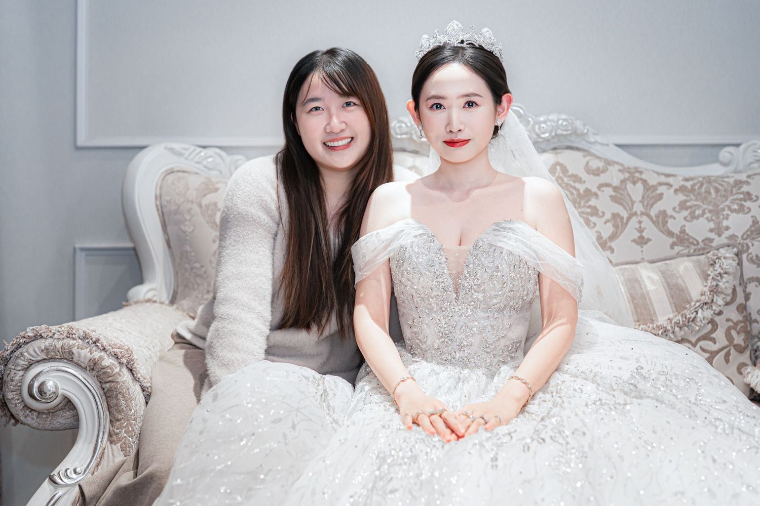 Two friends in elegant white wedding dresses pose together on a luxurious sofa in a bridal studio.