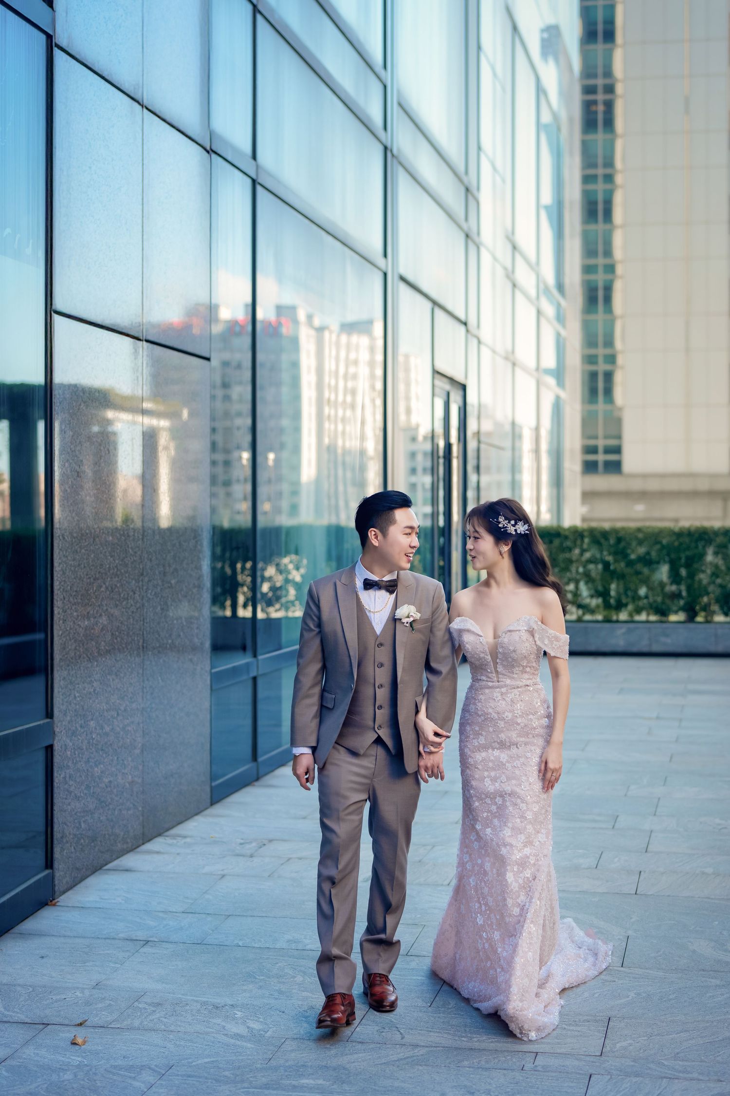 A couple in formal wedding attire walks hand in hand along a modern building exterior with glass windows.