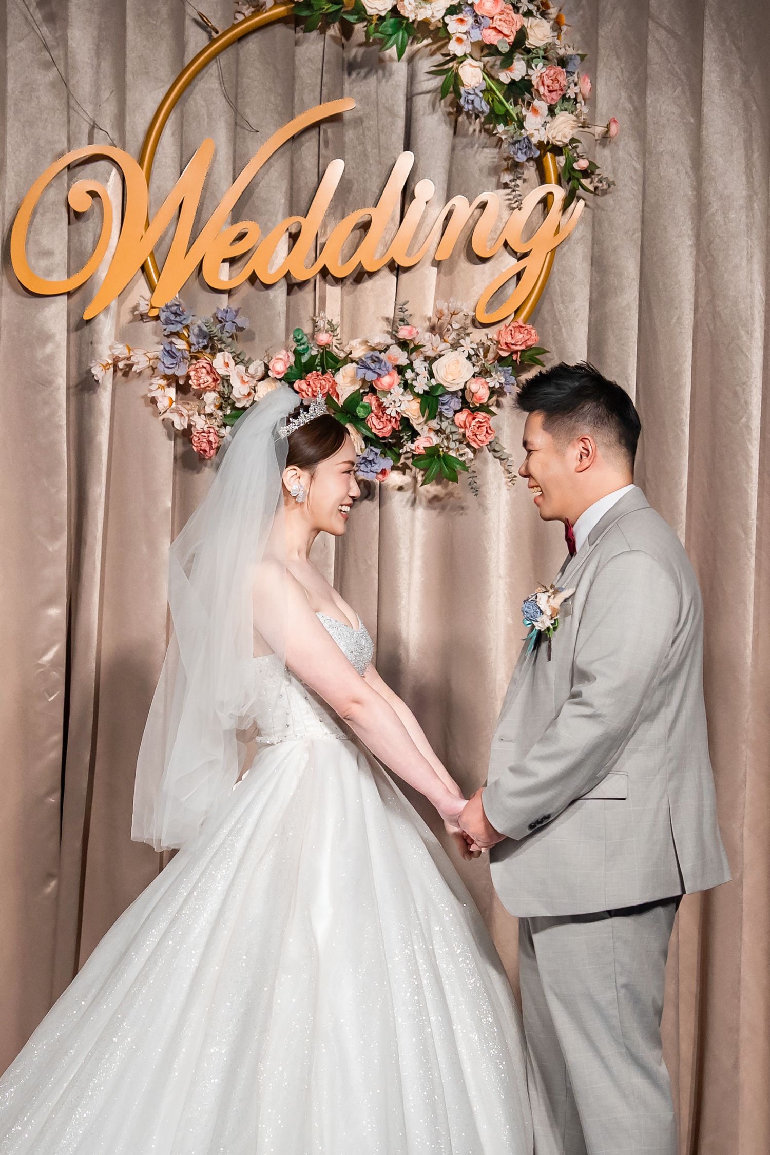A wedding couple poses against a floral backdrop with gold lettering in a romantic photo series.