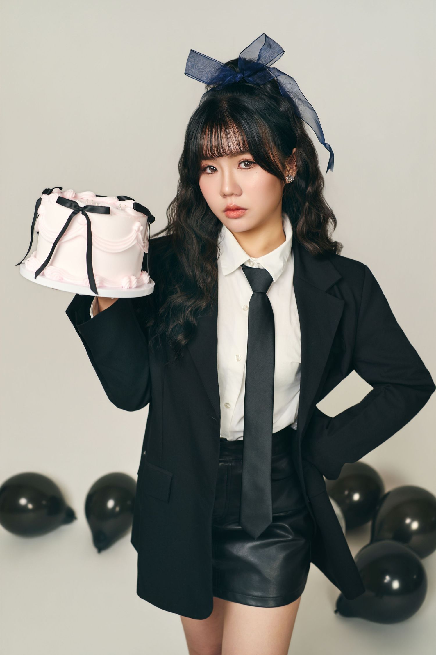A figure in a black blazer and white tie poses with various props against a light background with black balloons.