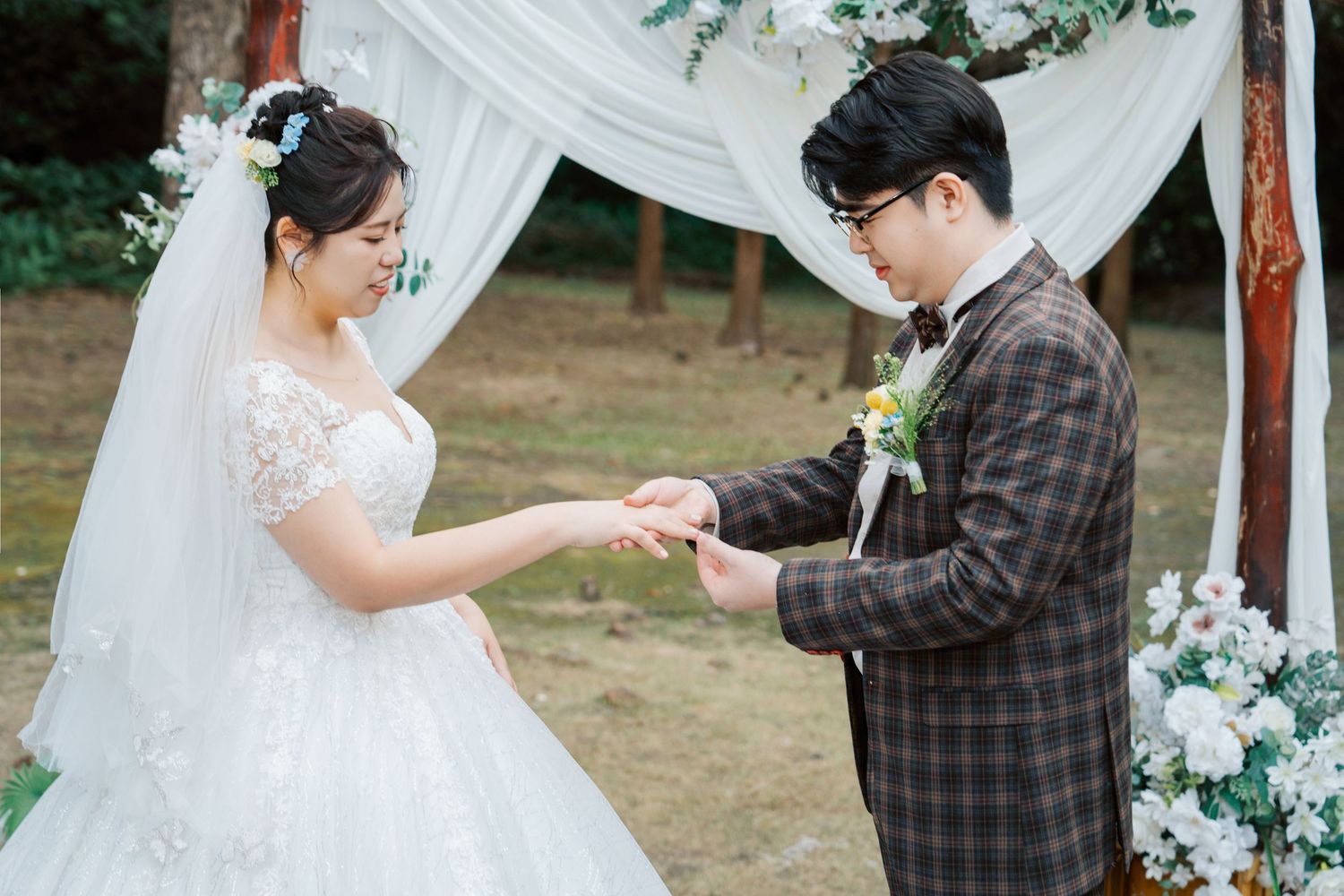 A couple exchanges rings during an outdoor wedding ceremony under a white draped arch decorated with greenery and flowers.