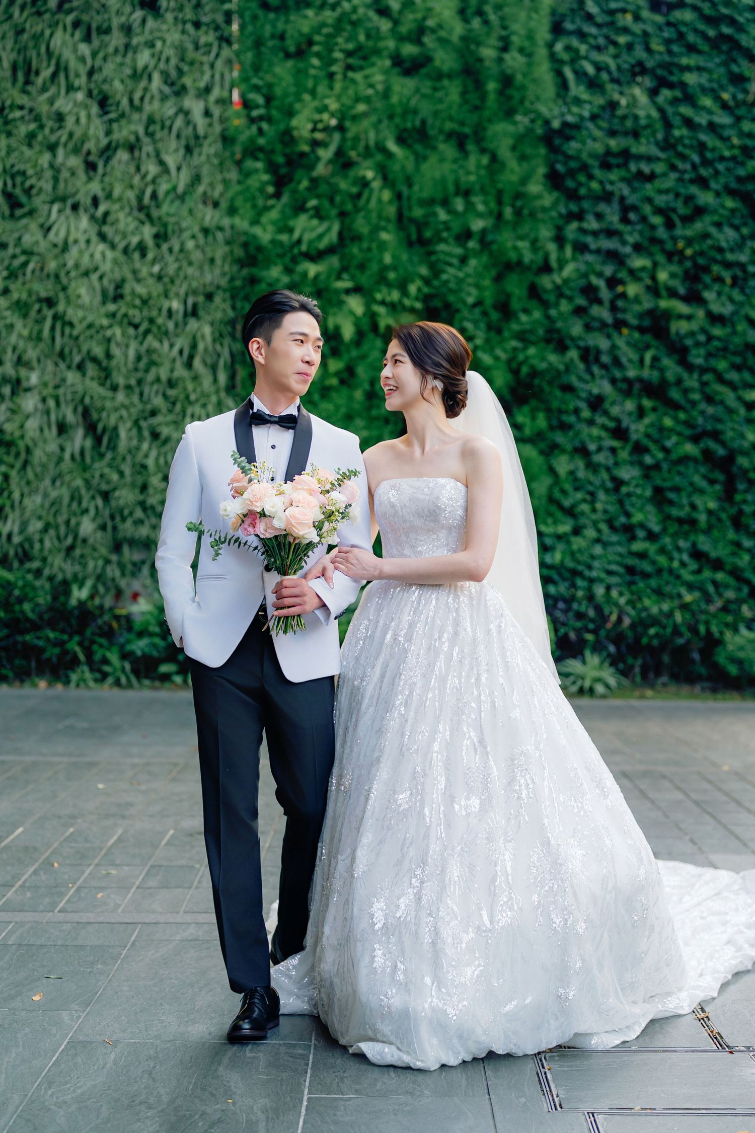A bride in a white lace gown and groom in light tuxedo pose together against a lush green hedge backdrop.