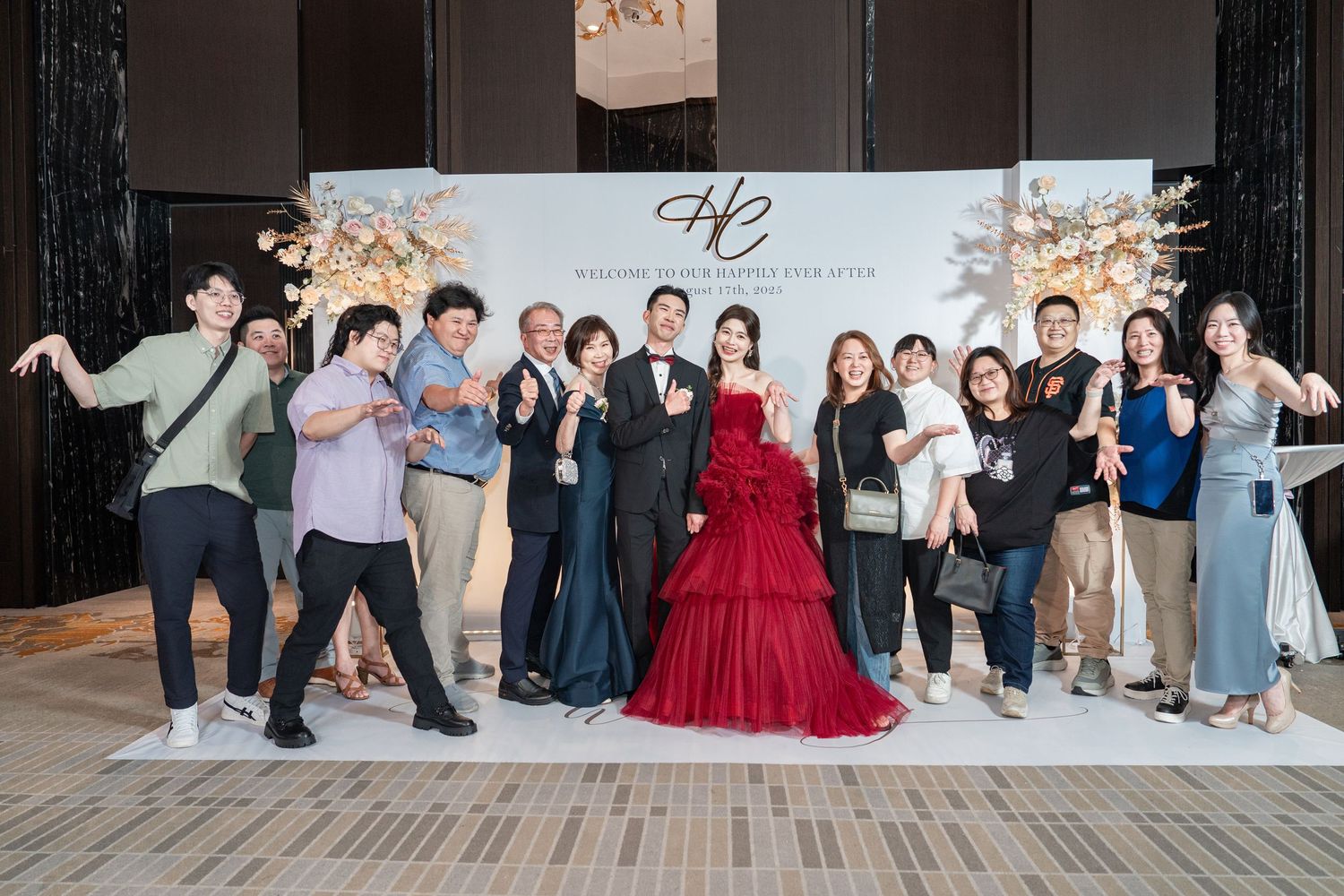 A group of party attendees pose together at a formal event with a red ballgown centerpiece against a decorated backdrop.