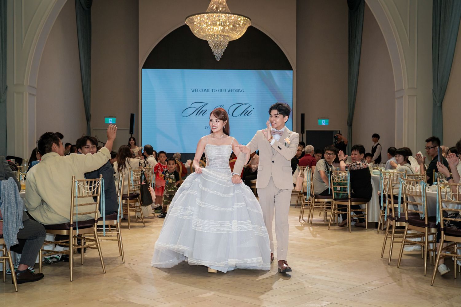 Bride in white ballgown and groom in beige suit walk together at elegant wedding reception with chandelier and blue backdrop.