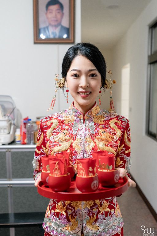 Traditional Chinese bride wearing a red qipao dress with gold embroidery holds a tray of tea cups for tea ceremony.