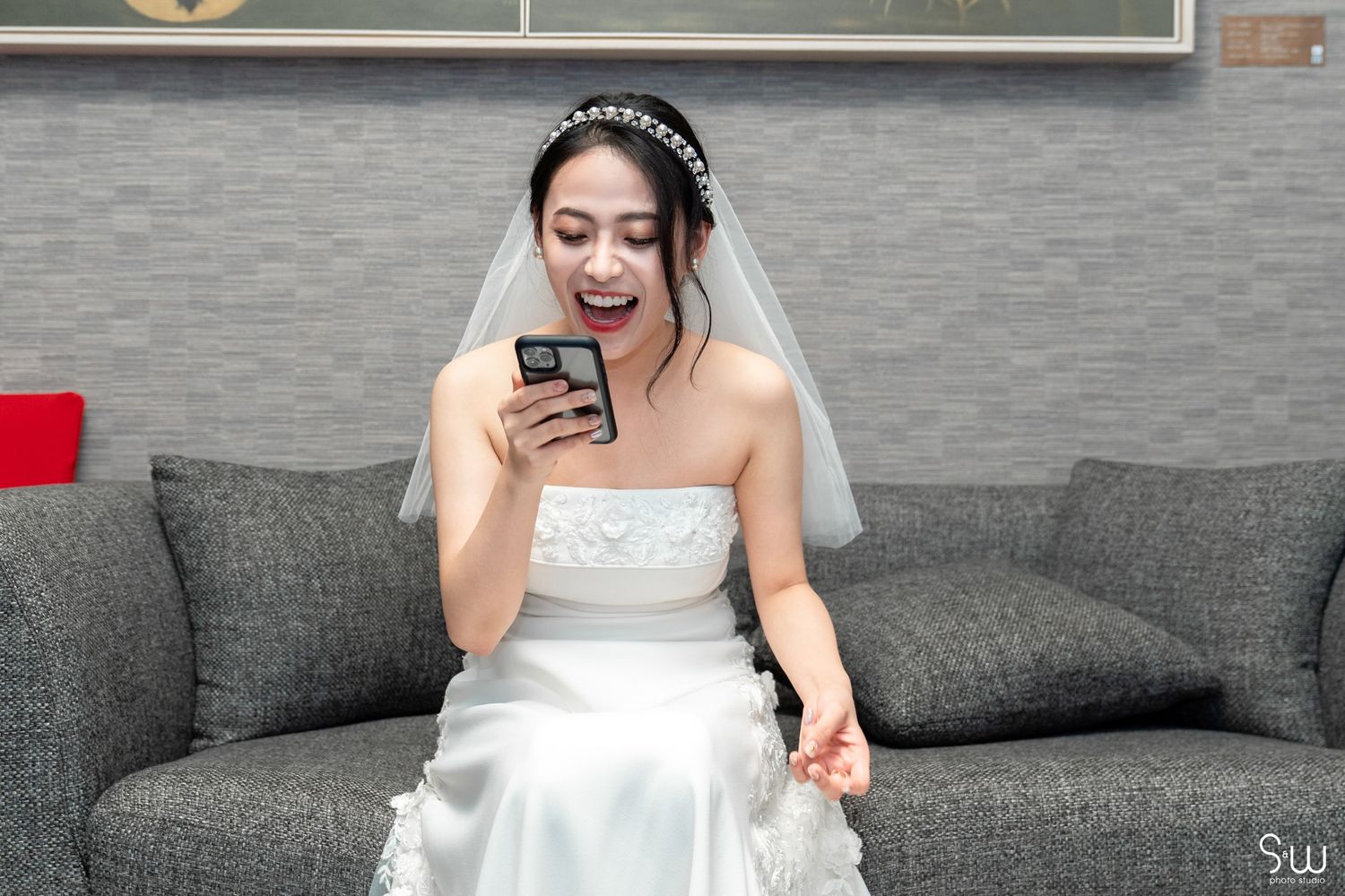 A bride in white dress sitting on gray couch reacts joyfully while looking at her phone.