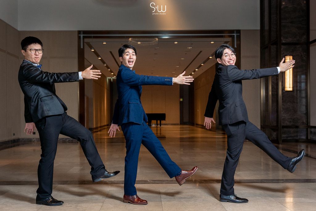 Three groomsmen in suits strike synchronized dramatic poses in a hotel hallway.