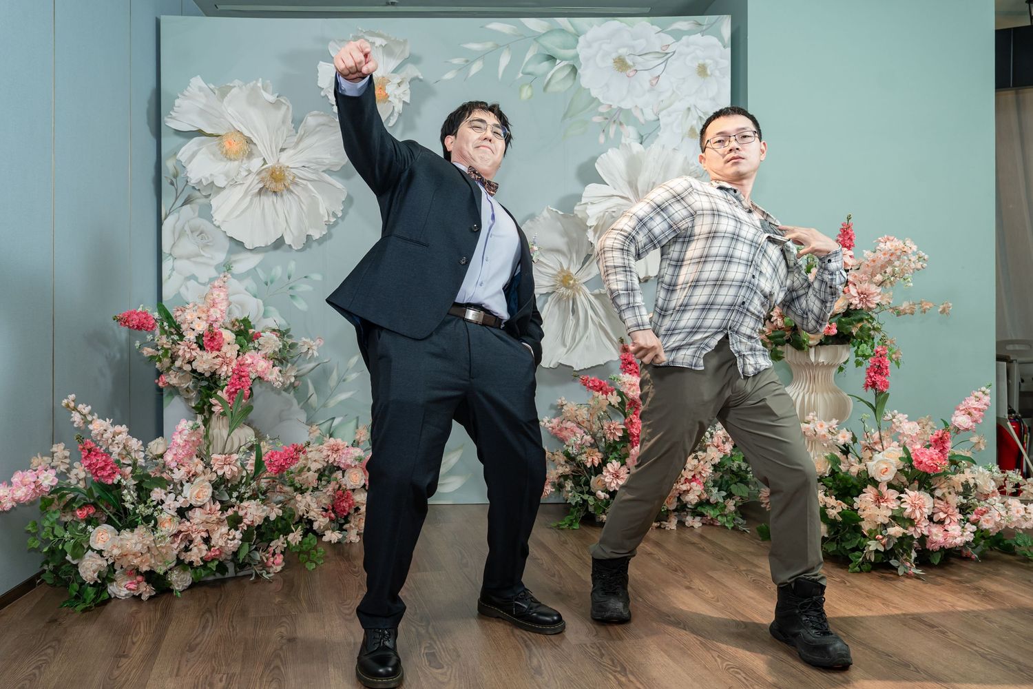Two dancers perform dynamic movements in a studio with pink floral decorations against a mint green wall.