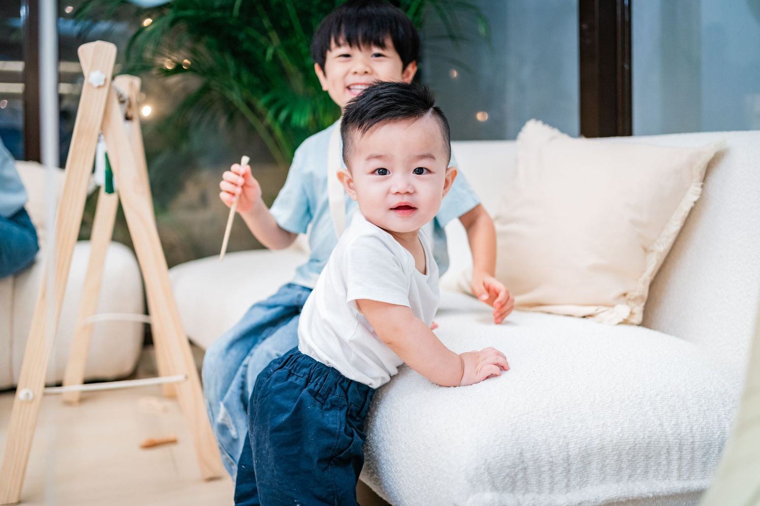 Baby in white clothing sits and plays on soft cushions near white furniture.