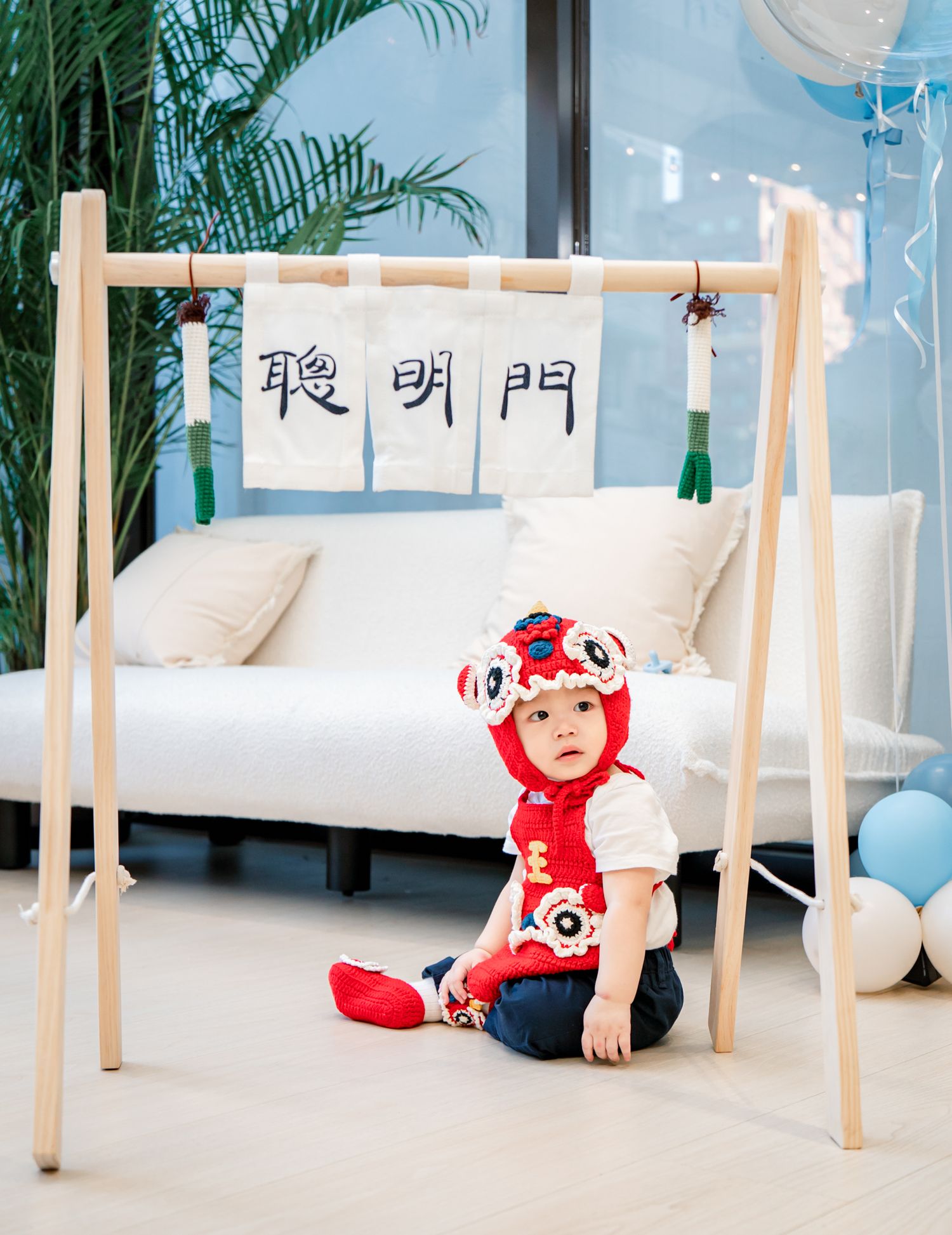Small child in red outfit crawls under wooden furniture in modern interior with palm plants and Chinese characters.