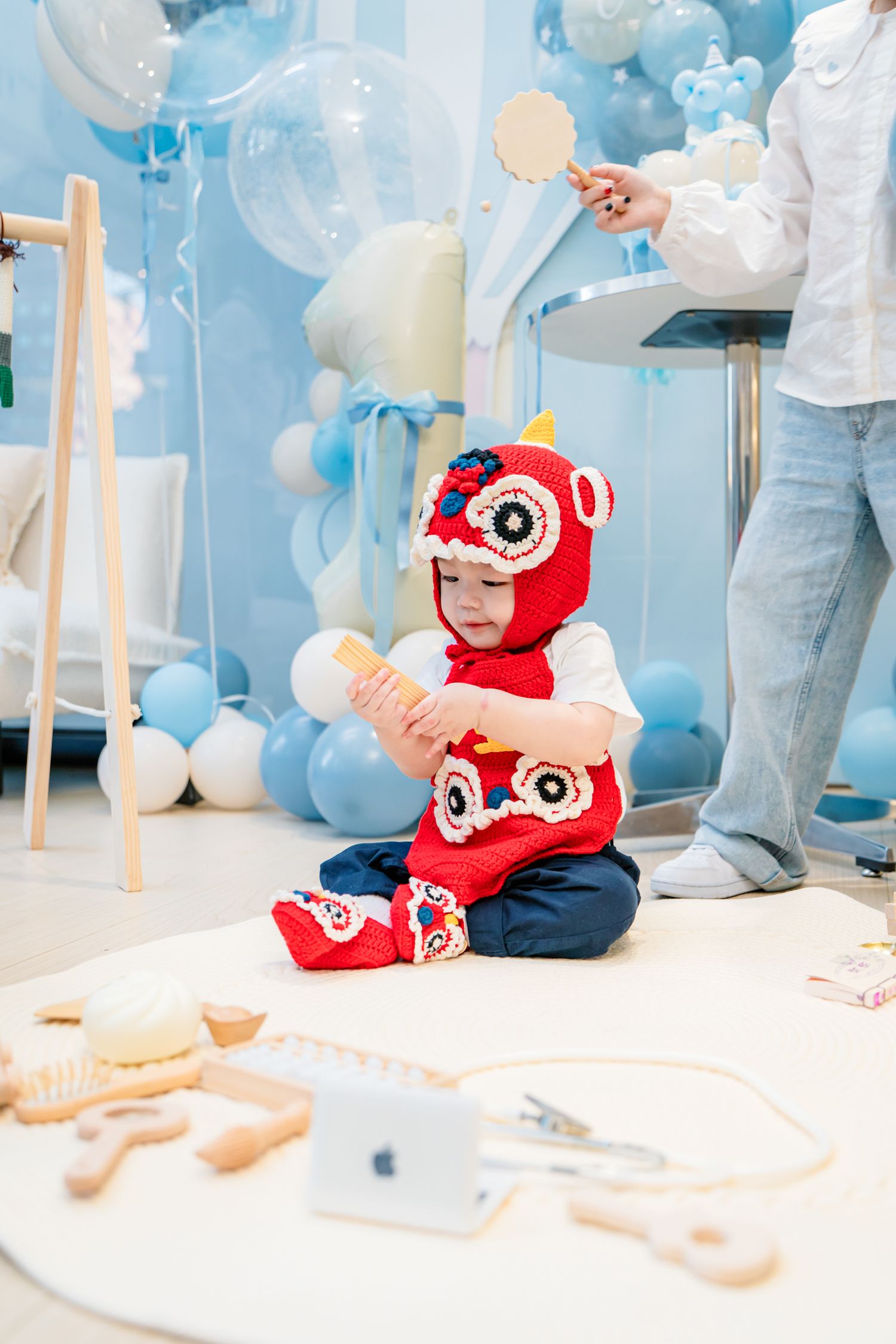 A baby in a red owl costume crawls on a white floor decorated with blue balloons and party decor.