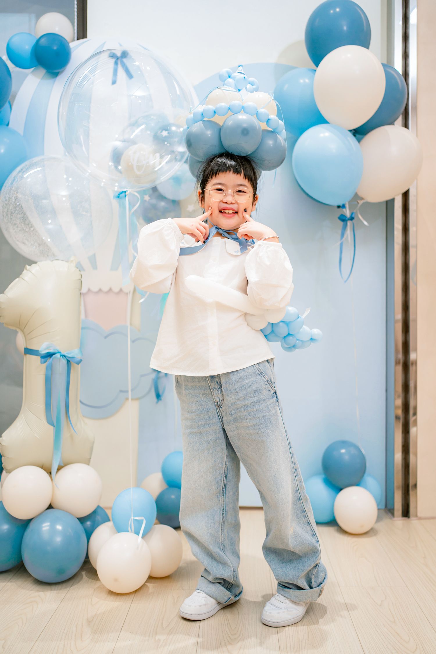 A child in a white sweater and jeans poses against a backdrop of blue and white balloons at a party celebration.