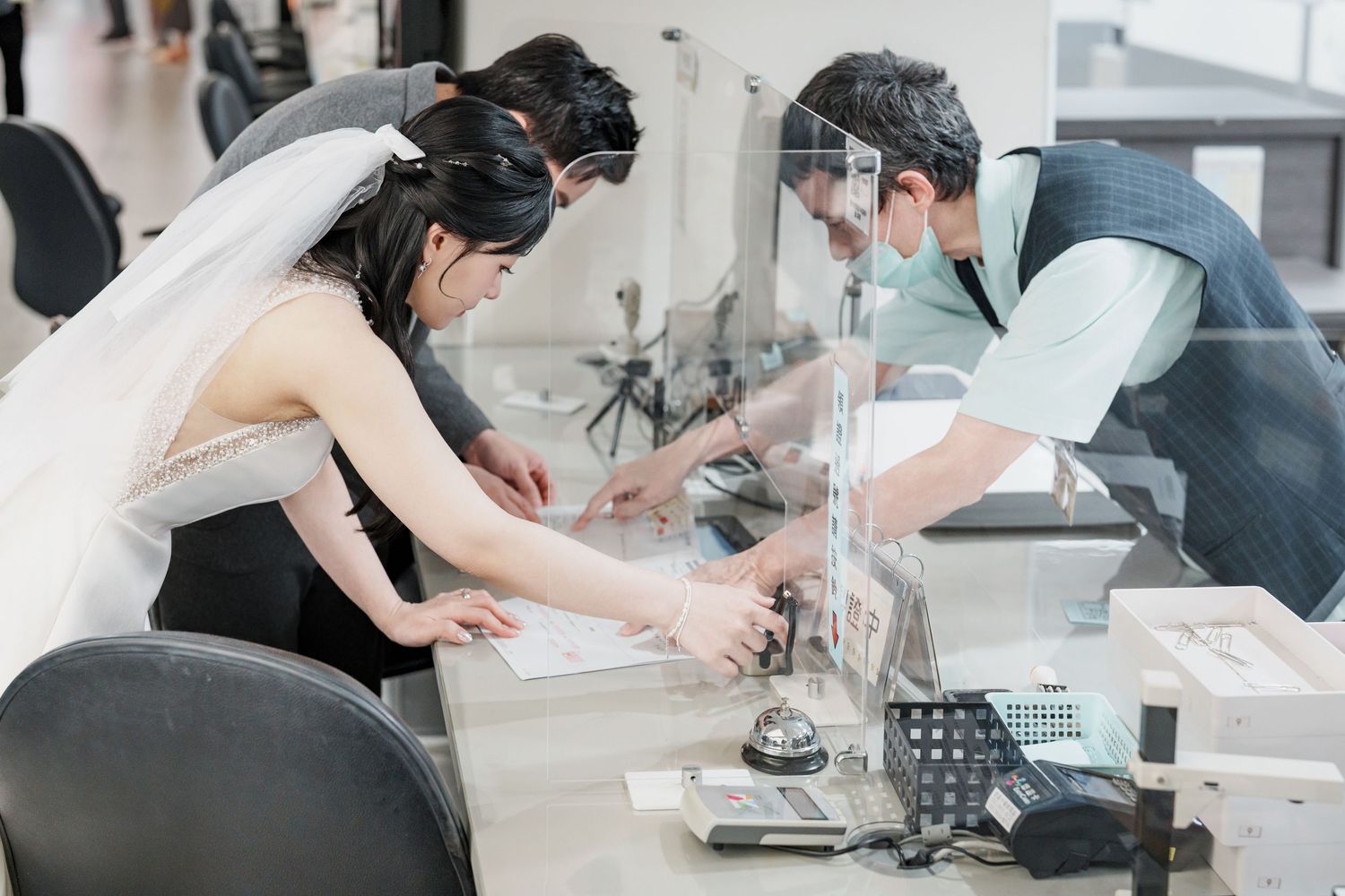 A customer and bank teller conduct a transaction through a protective glass barrier at a service counter.
