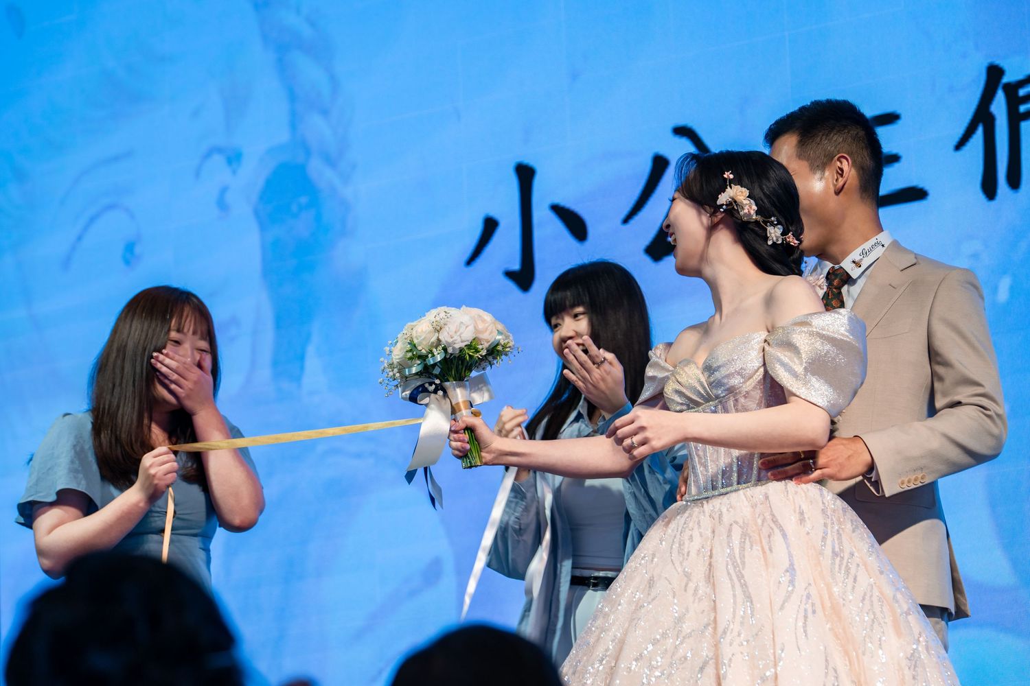 Bride in pink gown hands bouquet to emotional guests on stage at a wedding ceremony.