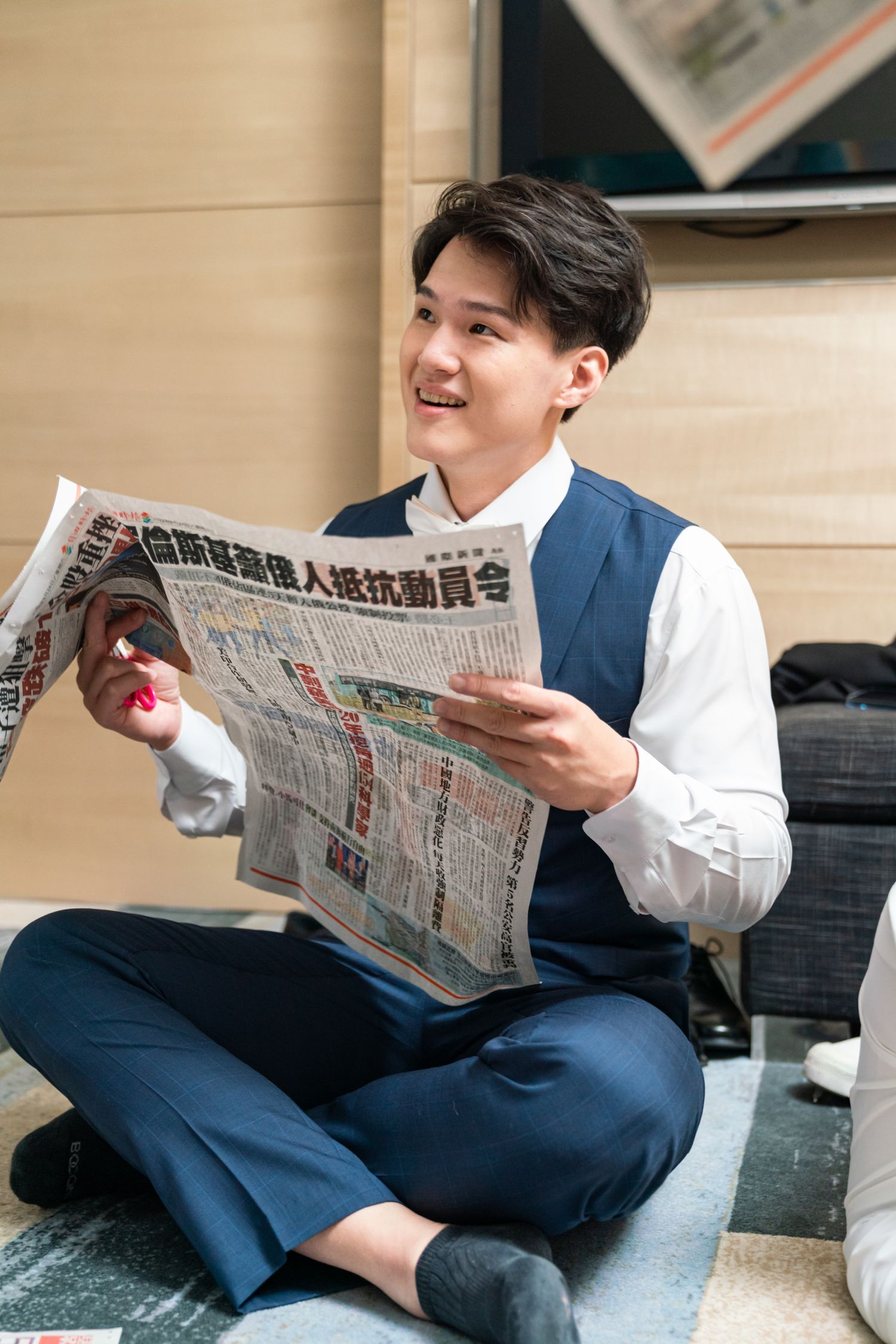 A smiling person in business attire sits on the floor reading a Chinese newspaper with interest.