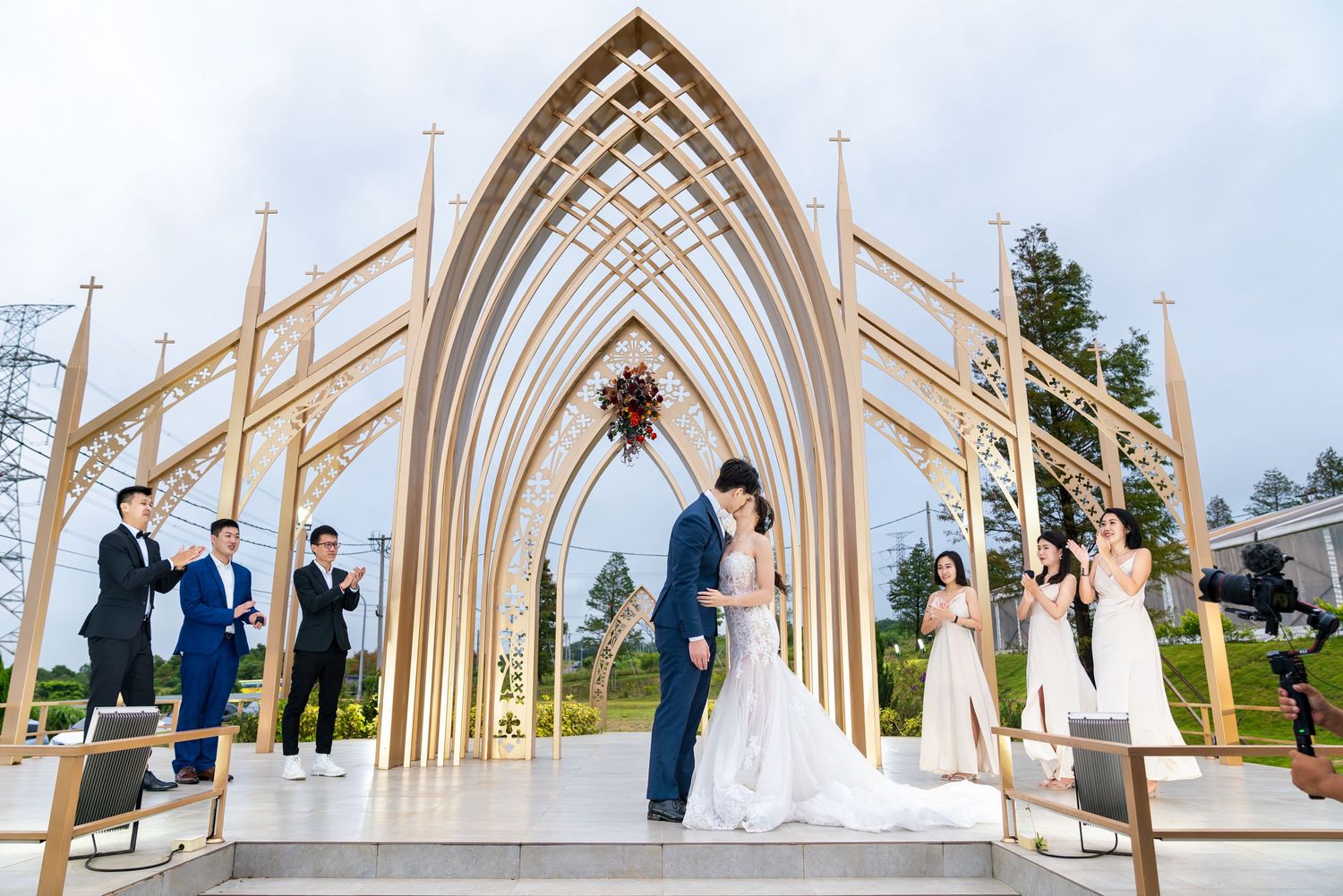 A wedding ceremony takes place under an elegant arched white chapel pavilion with groomsmen and bridesmaids in attendance.
